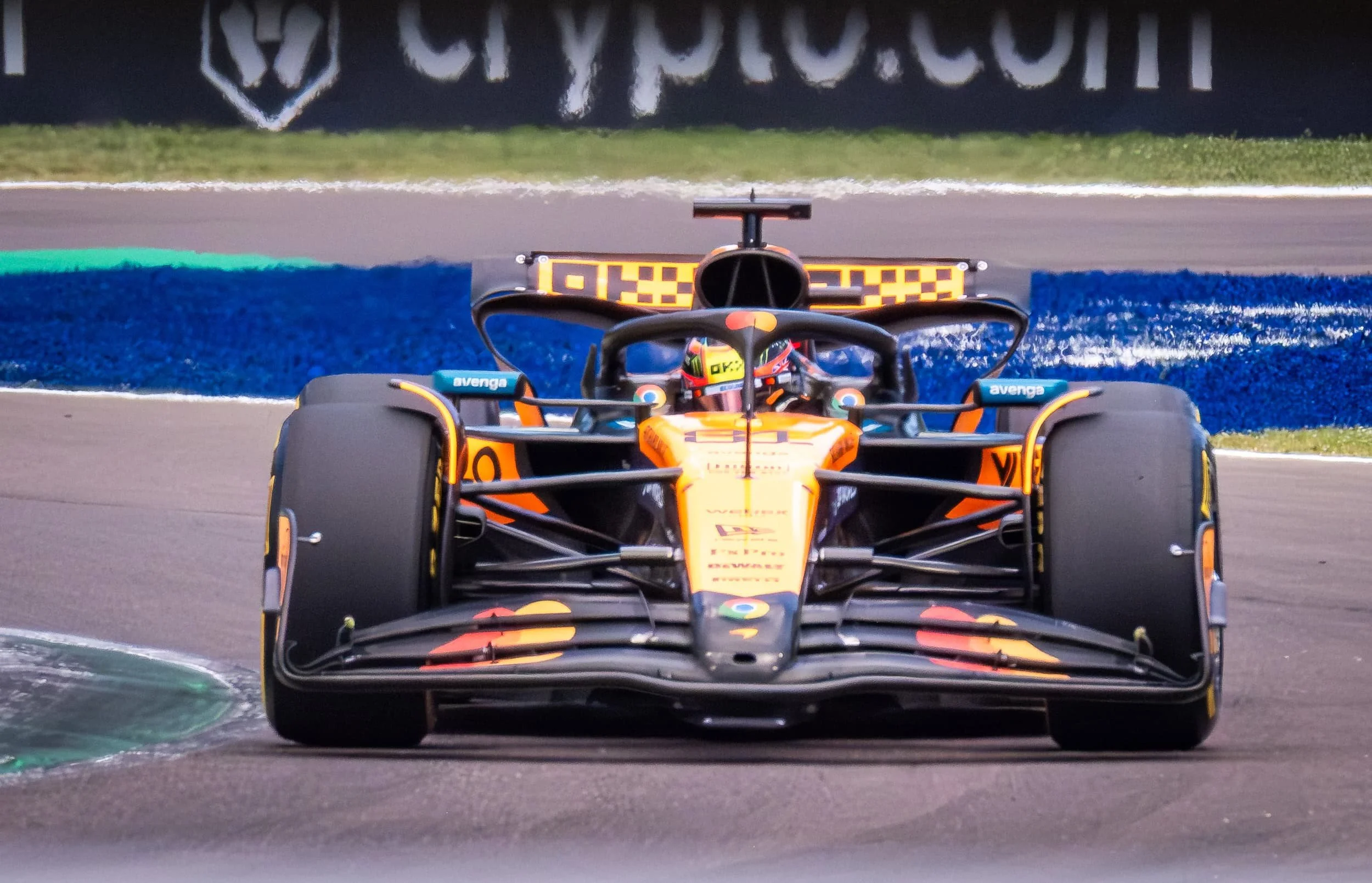 A racing car on a track, seen from the front, with bright orange, black, and yellow livery, and a driver wearing a helmet. The tire marks indicate high-speed movement.