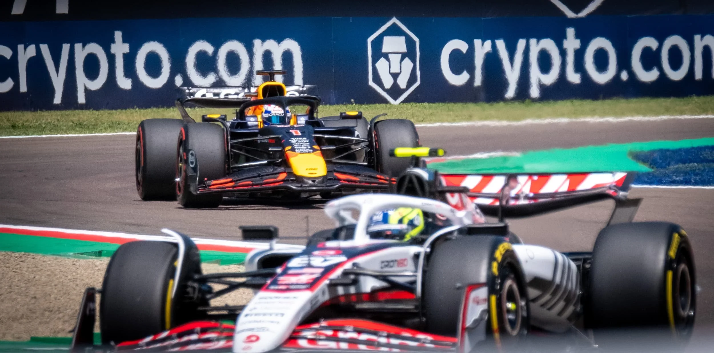 Race cars on a track during a race, with one car in the foreground and another behind, featuring a blue advertisement banner in the background.