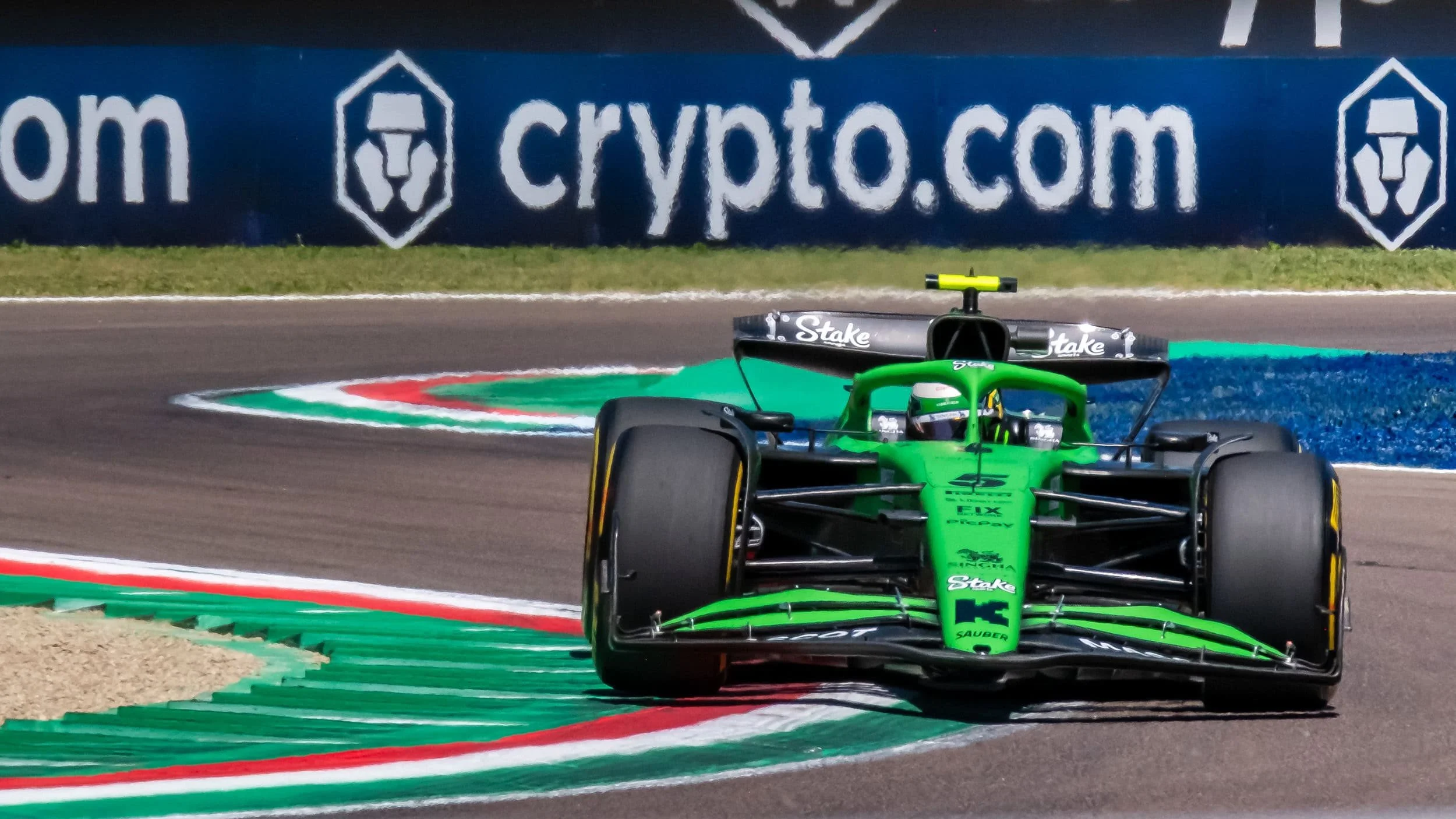 Green racing car on a race track with a driver wearing a helmet, near a curb painted in green, white, red, and blue, with a blue advertising banner in the background.