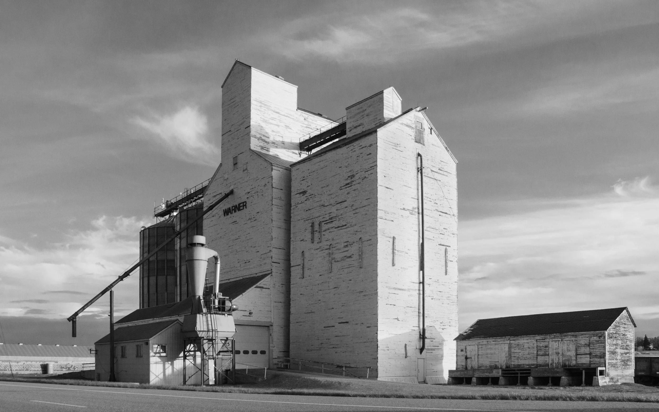 Black and white photograph of a historic grain elevator in Warner, Alberta, capturing the stark geometry, weathered textures, and quiet presence of rural prairie architecture under a wide open sky.