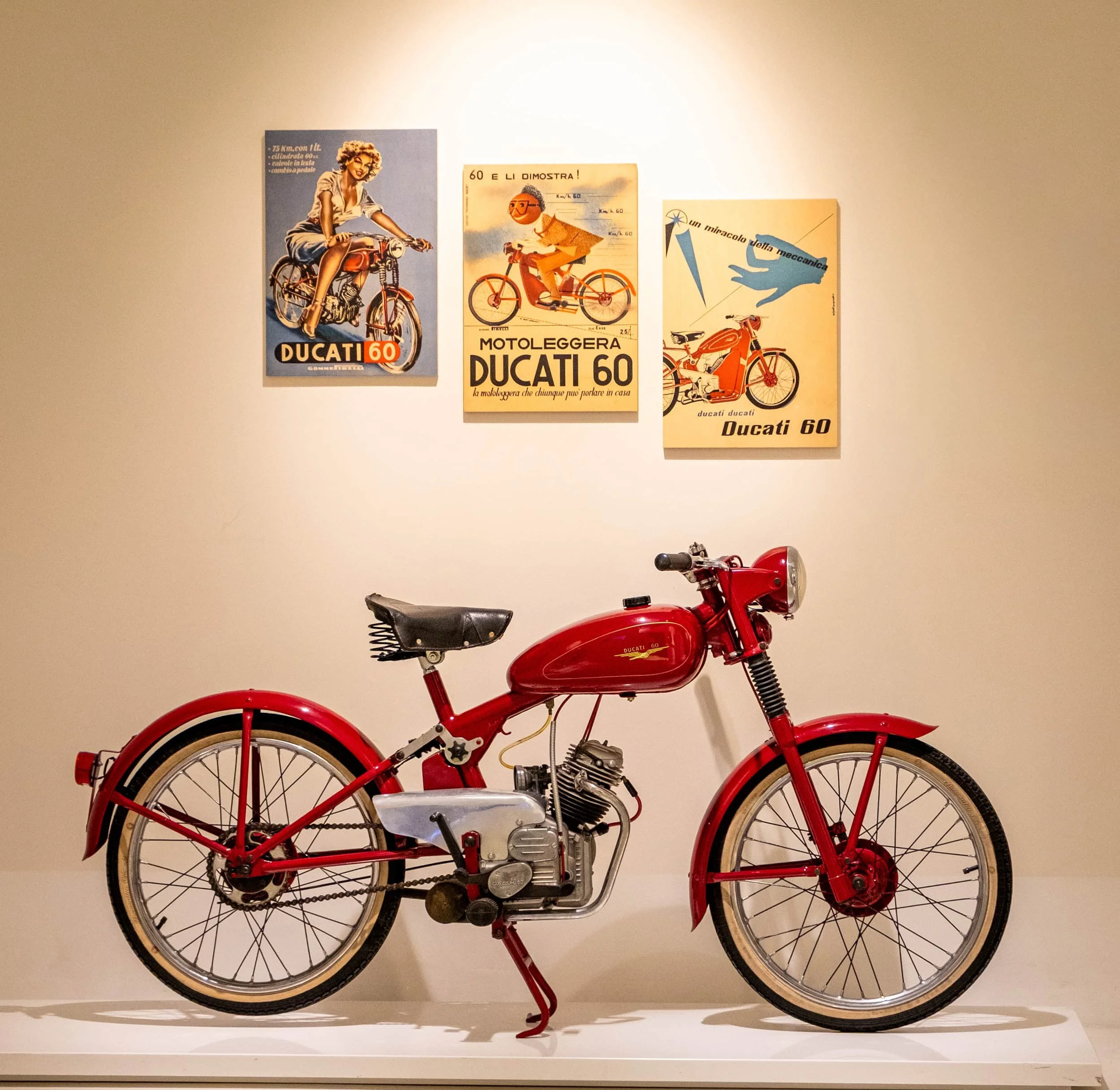 The Ducati Cucciolo, a motorized bicycle displayed at the Ducati Museum in Bologna, Italy, with classic Ducati advertising posters mounted on the wall above.
