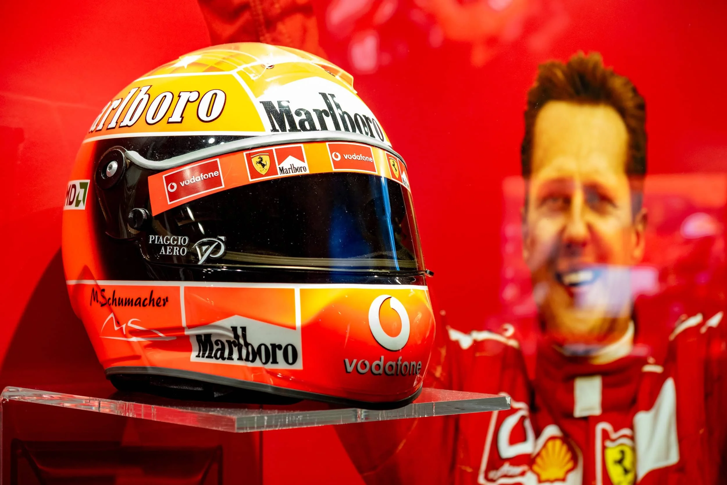 Michael Schumacher’s Ferrari F1 racing helmet displayed at the Ferrari Museum in Maranello, Italy, with a portrait of Schumacher in the background.
