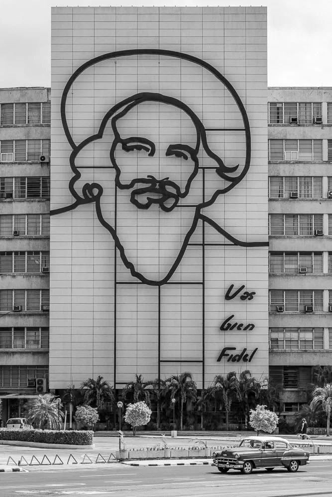 Black and white image of the Ministry of Informatics and Communications building with Camilo Cienfuegos mural and “Vas bien, Fidel” slogan.