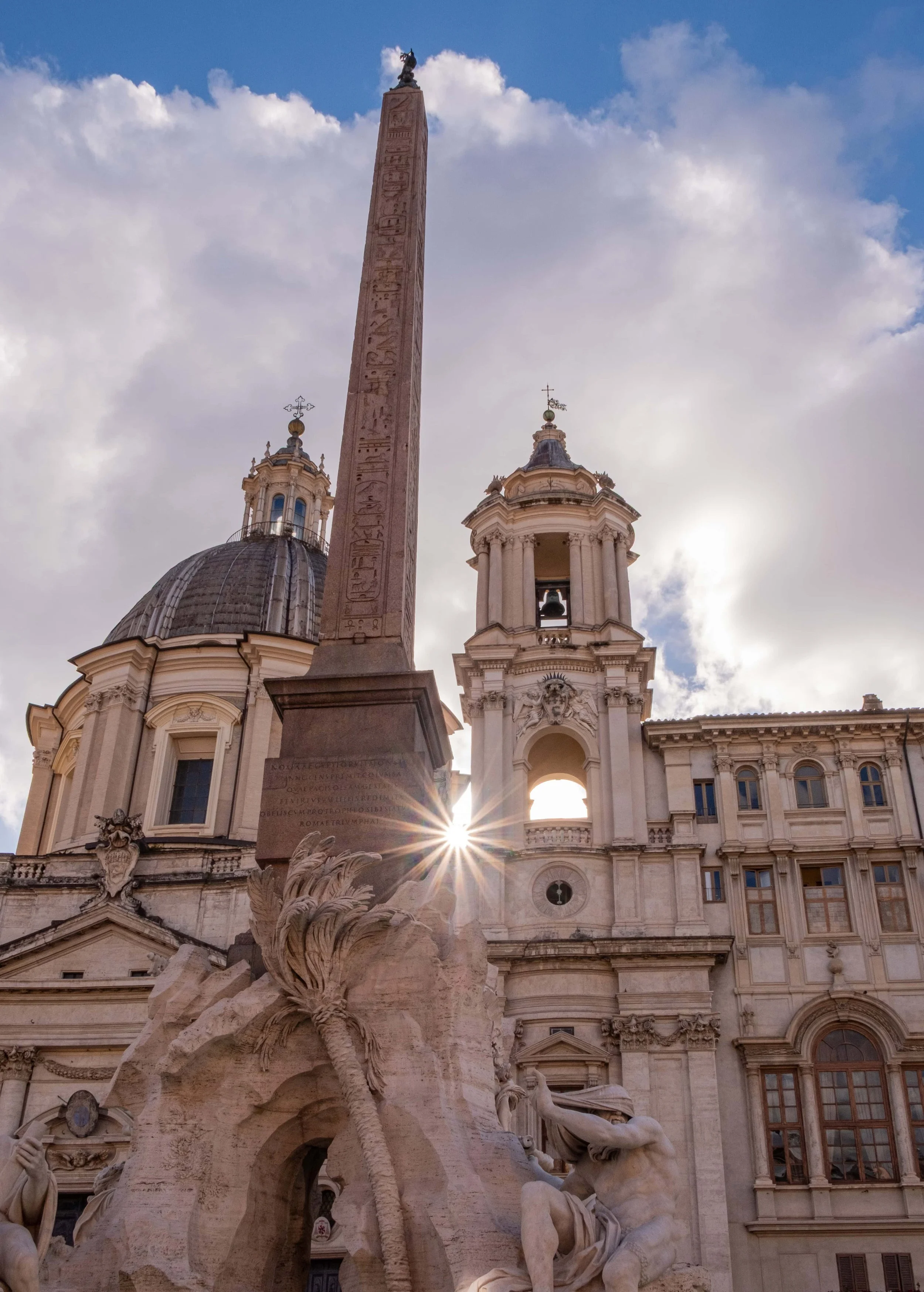 A dramatic view in Piazza Navona, Rome, where the obelisk of the Fountain of the Four Rivers rises in the foreground while the Baroque façade and dome of Sant’Agnese in Agone stand behind it. A sunburst flares through the narrow space between the str