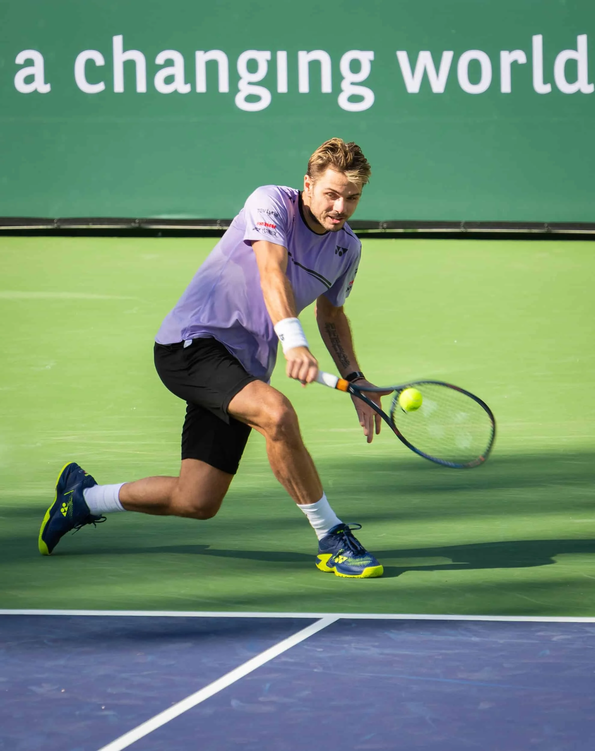 tan (the man), gets low to hit a one-handed backhand at  Indian Wells, CA. He won his match against Daniel Evans 6-7, 6-3,6-3.