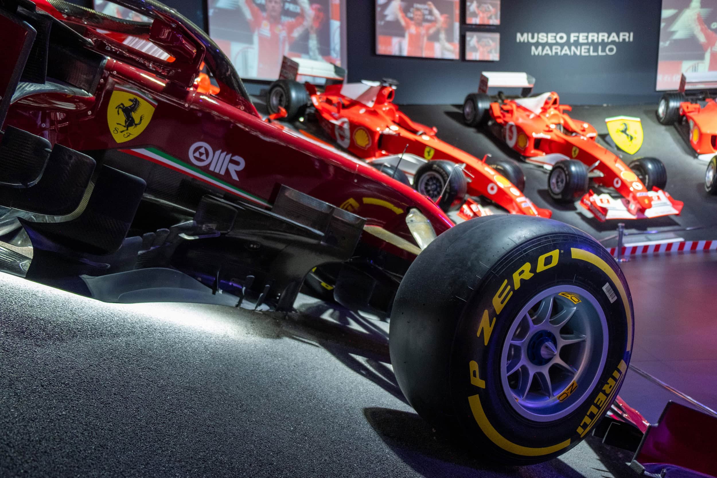 Close-up of a modern Ferrari Formula 1 race car with Pirelli tires displayed inside the Ferrari Museum in Maranello, Italy, with historic Ferrari F1 cars visible in the background.