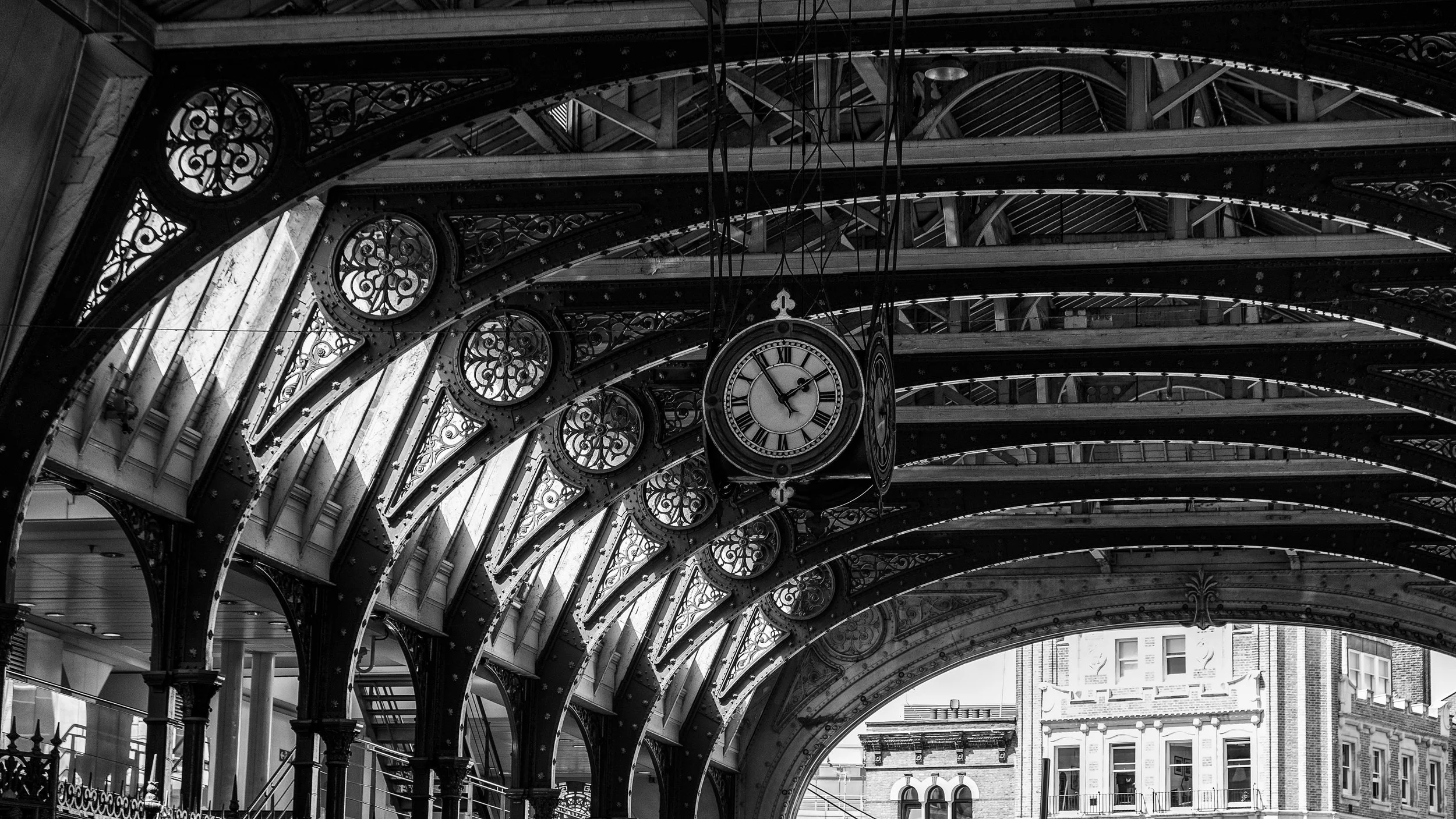The historic Victorian ceiling of Old Spitalfields Market in London showcases an intricate lattice of iron and glass arching overhead. Suspended at its center, a vintage clock hangs beneath the softly filtered daylight, highlighting the architectural
