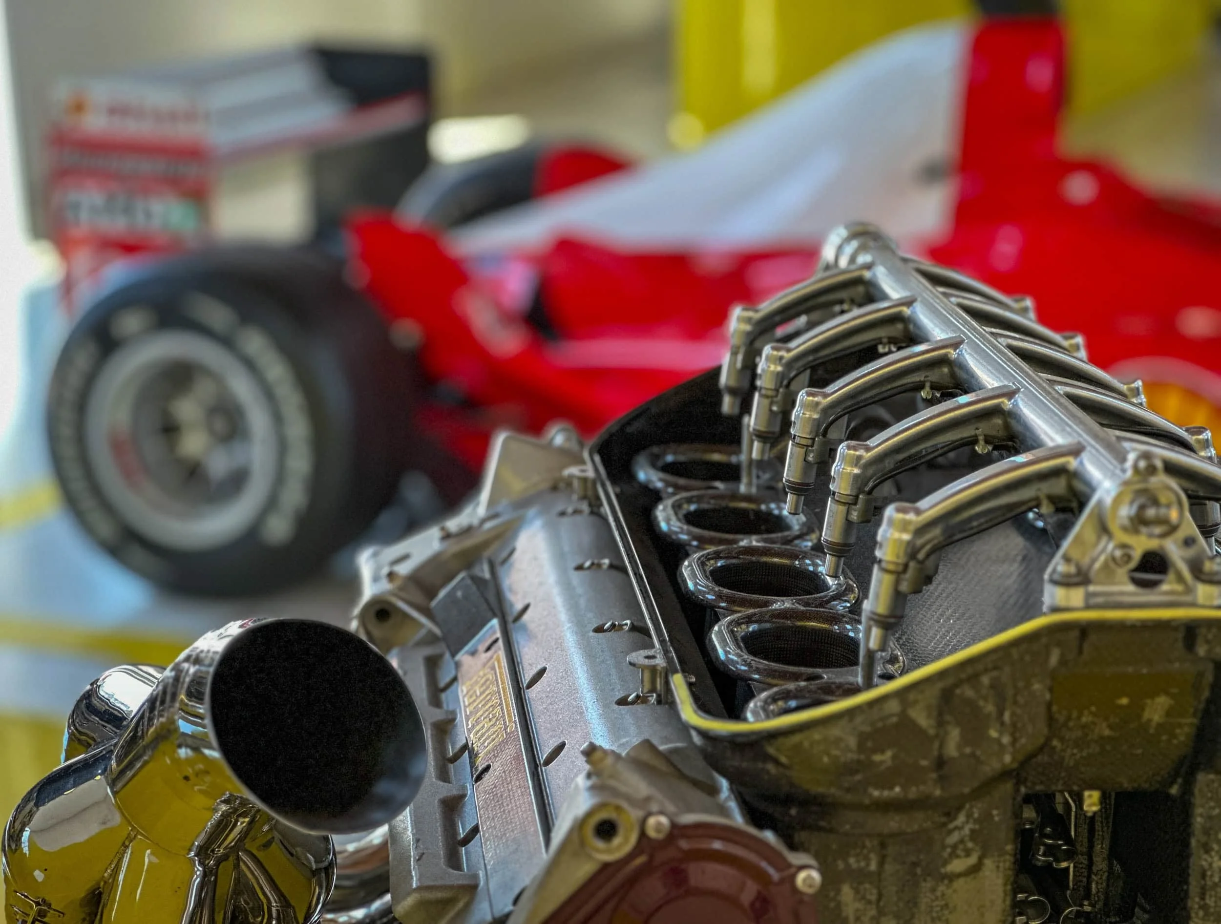 lose-up of a Ferrari Formula 1 V10 engine display with intake trumpets in sharp focus and a red Ferrari F1 race car blurred in the background at the Enzo Ferrari Museum in Modena, Italy.