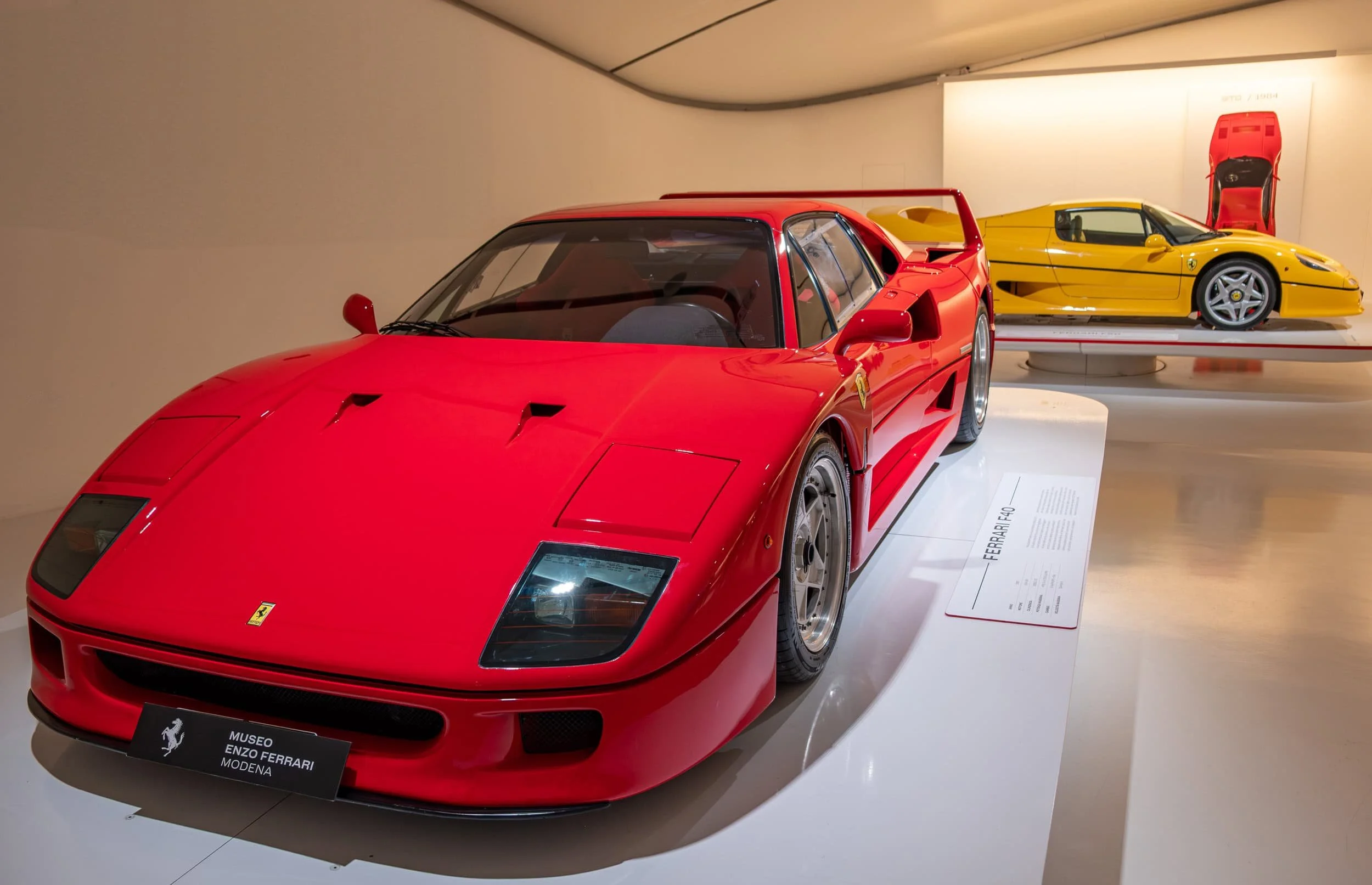 Red Ferrari F40 displayed at the Enzo Ferrari Museum in Modena, Italy, with a yellow Ferrari F50 visible in the background inside the Enzo Ferrari Museum