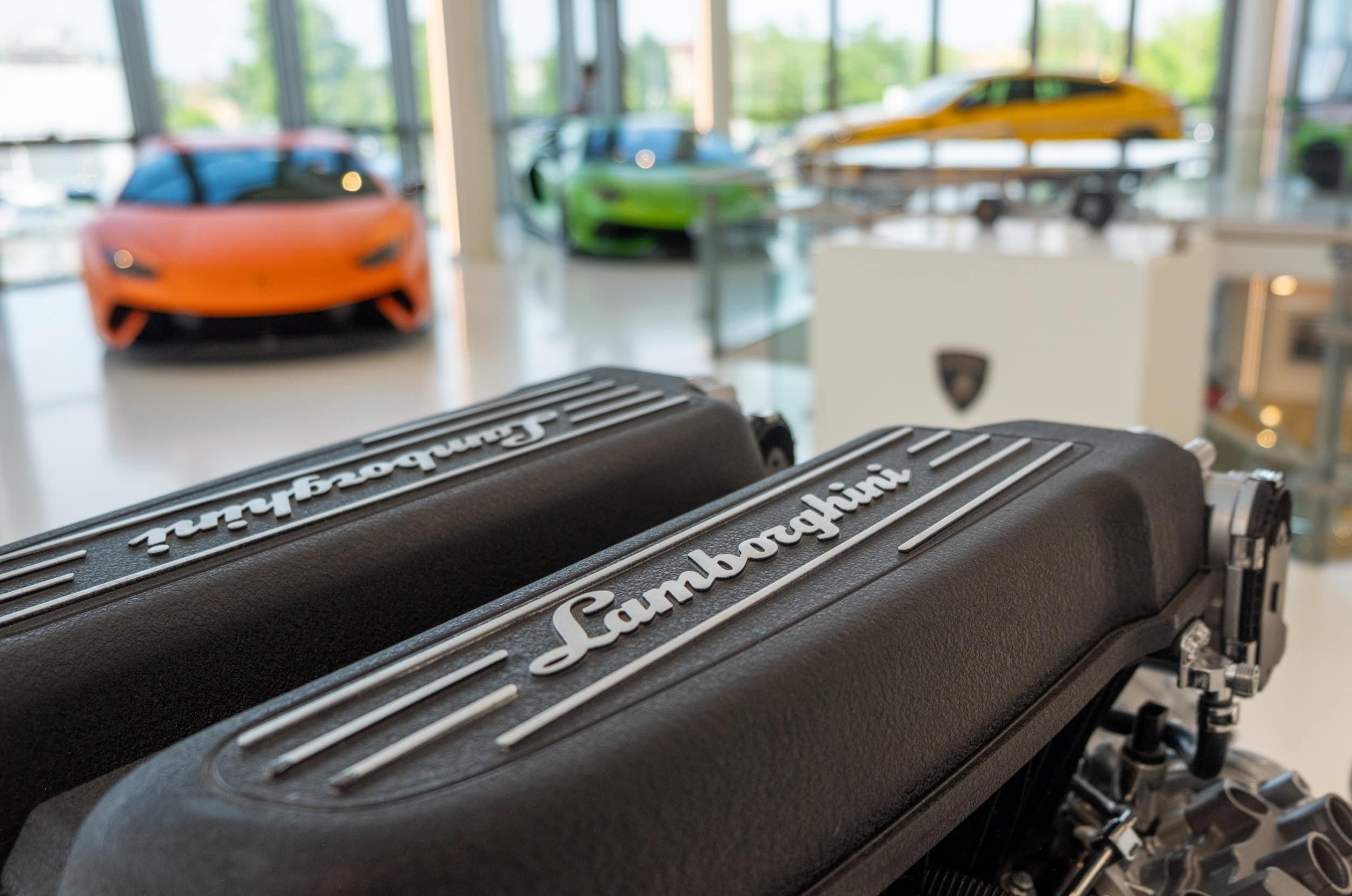 A close-up view of a Lamborghini valve cover contrasting with the softly blurred backdrop of some of the newer models (Huracan and Urus).