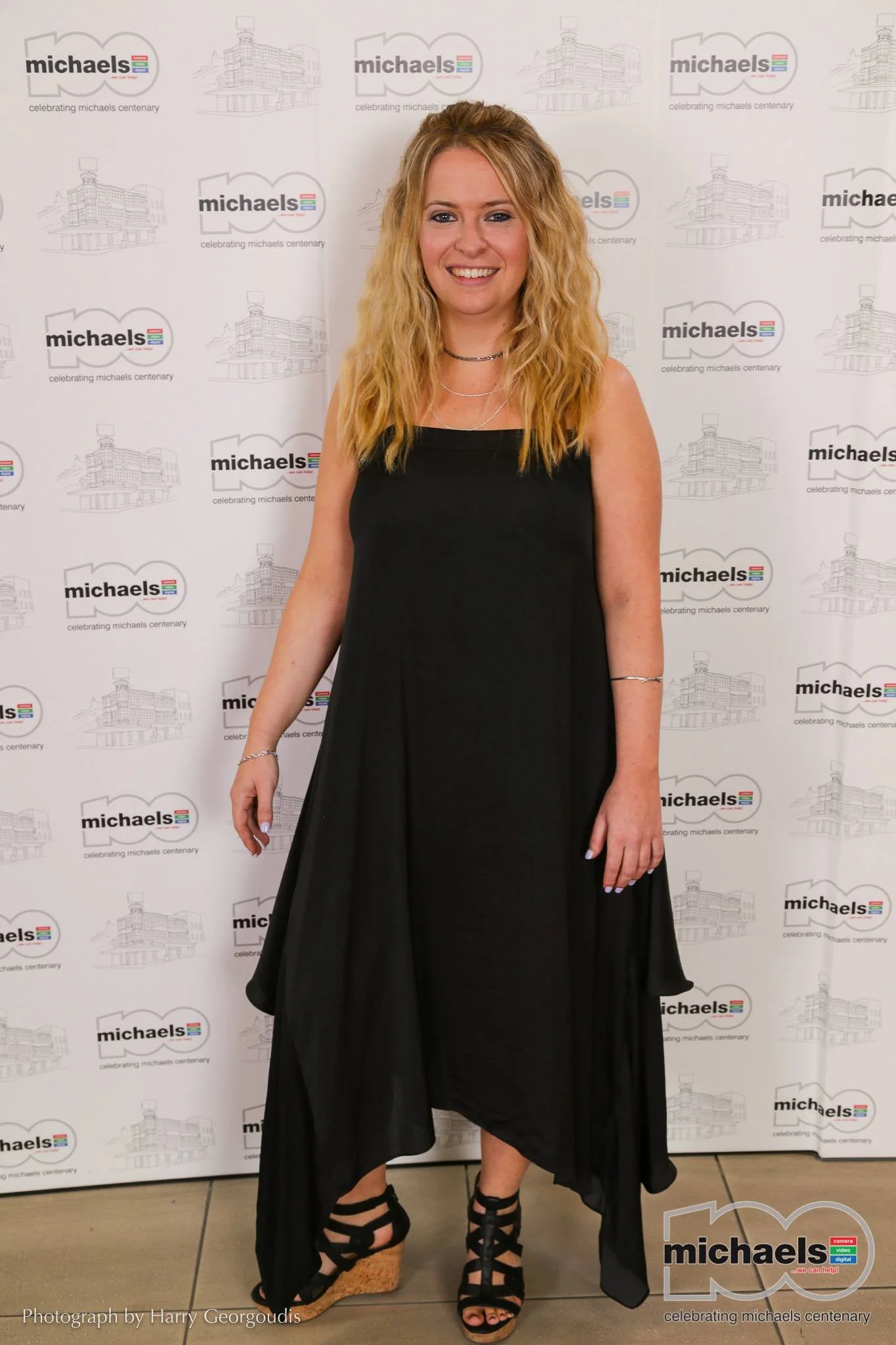 Carly Michael standing in front of a media wall dressed elegantly at the michaels centenary gala event