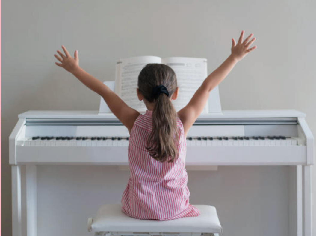 A young girl with brown hair tied back, wearing a red and white striped dress, sits at a white piano with her arms raised in celebration or excitement while looking at an open sheet music.