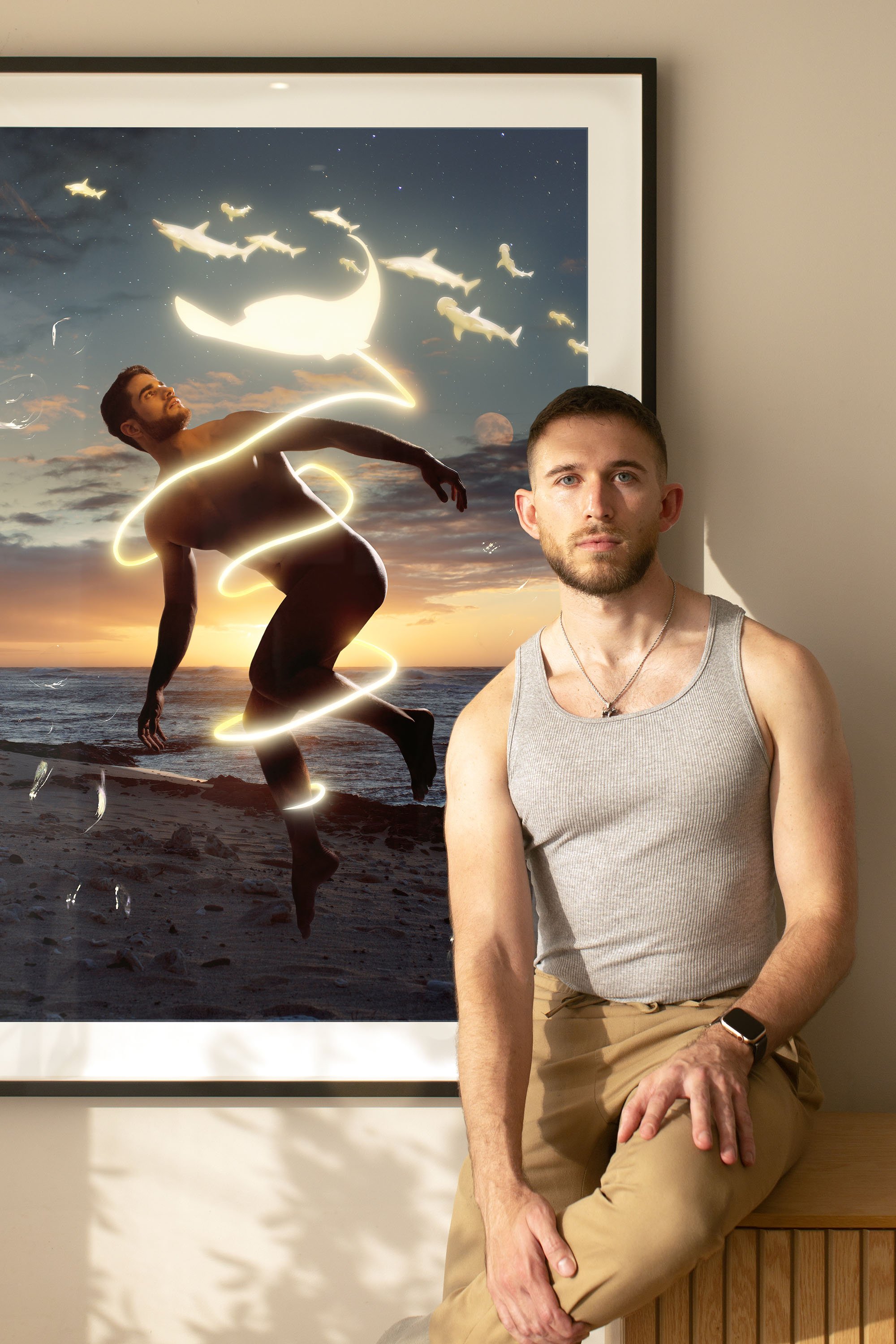 A man sitting next to a window with a framed painting of a beach sunset scene, featuring a woman with glowing lines circling her body and a moon and fish in the sky.