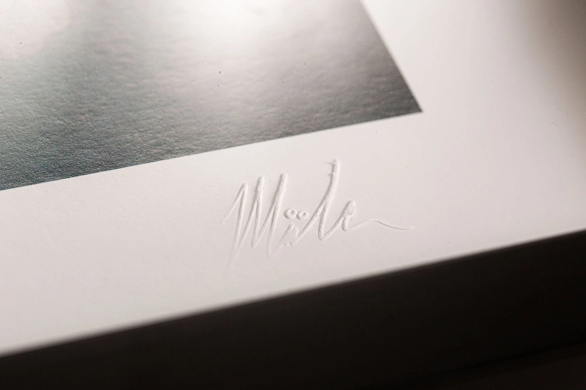 Close-up of embossed signature on white paper, with a textured metallic gray surface in the top left corner.