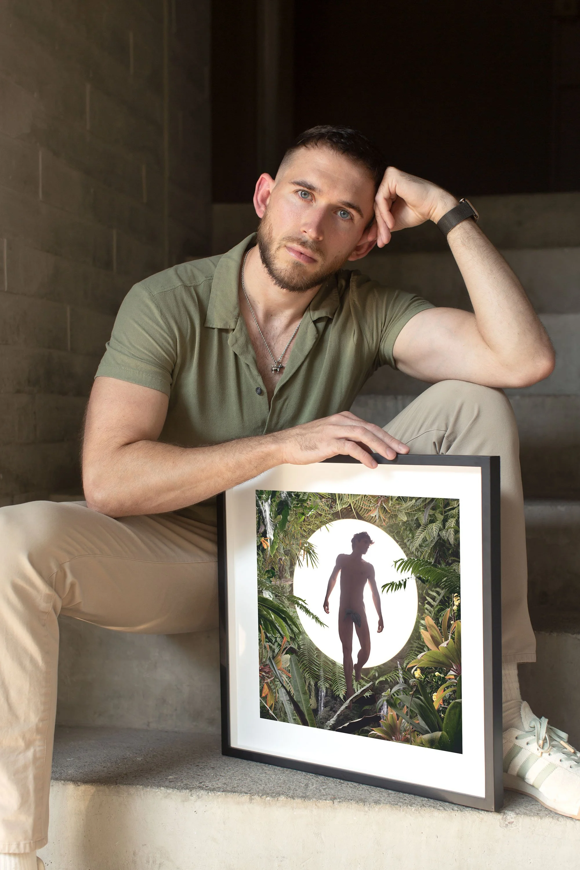 A young man with short dark hair, beard, and light blue eyes sitting on steps with hand on his head, holding a framed artwork of a silhouette of a nude man standing among lush tropical plants and leaves.