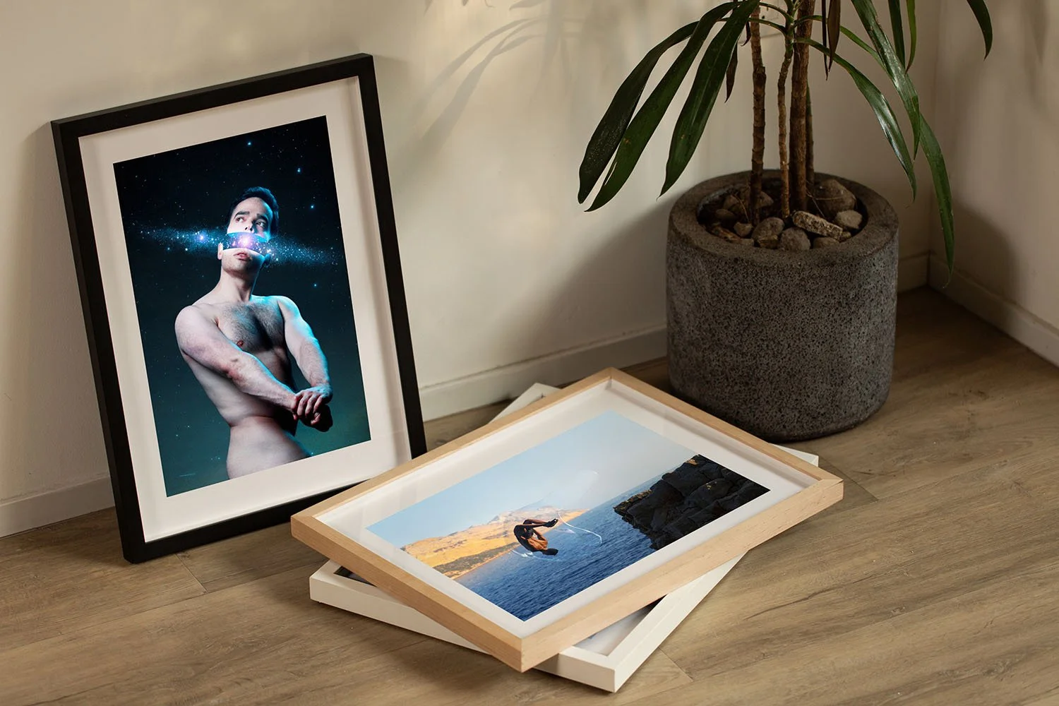 Stack of framed photographs on a wooden floor next to a potted plant with long green leaves. One photo features a shirtless man with arms crossed, against a starry background. The other photo shows a person diving into the water near a rocky shoreline.