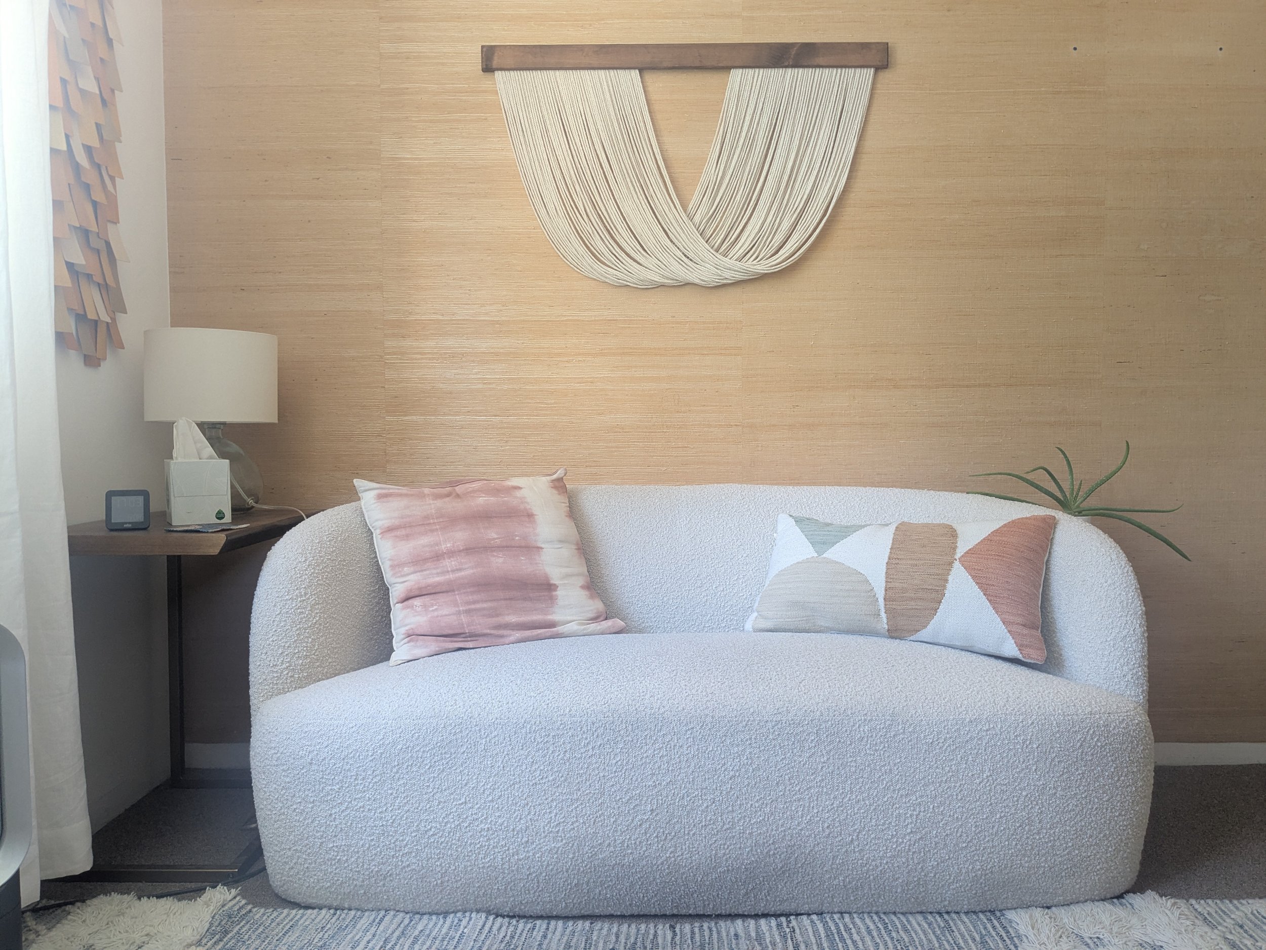 A cozy white textured sofa, pink and patterned throw pillows, a wooden side table with a digital clock, tissue box, and lamp, a beige textured wall with a modern fabric art piece, a window with curtains, a small plant in the bottom right corner, and a soft cream-colored rug.