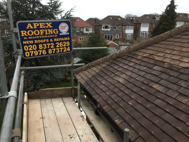 View from a rooftop under construction, with scaffolding and a sign for Apex Roofing & Maintenance advertising services for new roofs and repairs, with contact phone numbers, in a neighborhood with residential houses in the background.