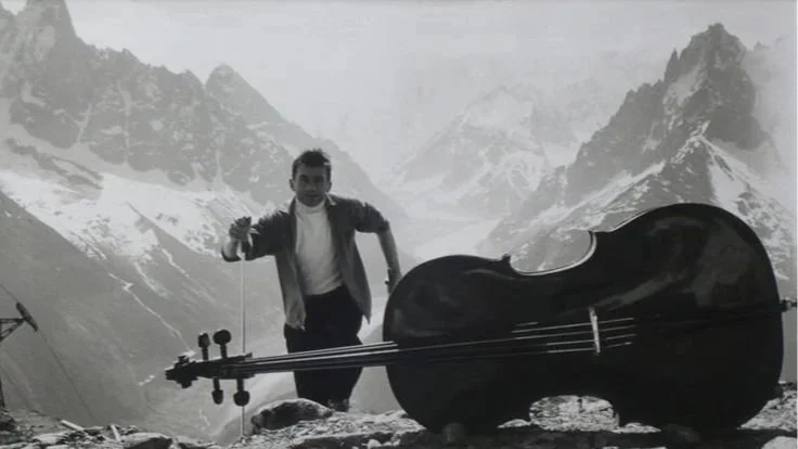 Black-and-white image of a man standing beside a cello against a snowy mountain landscape