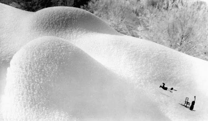 Black-and-white photo of a skier tumbling down a steep powder-covered snowy slope near ski equipment.