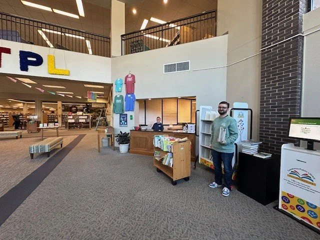 large two-story library lobby with staff member standing near checkout machine on the right
