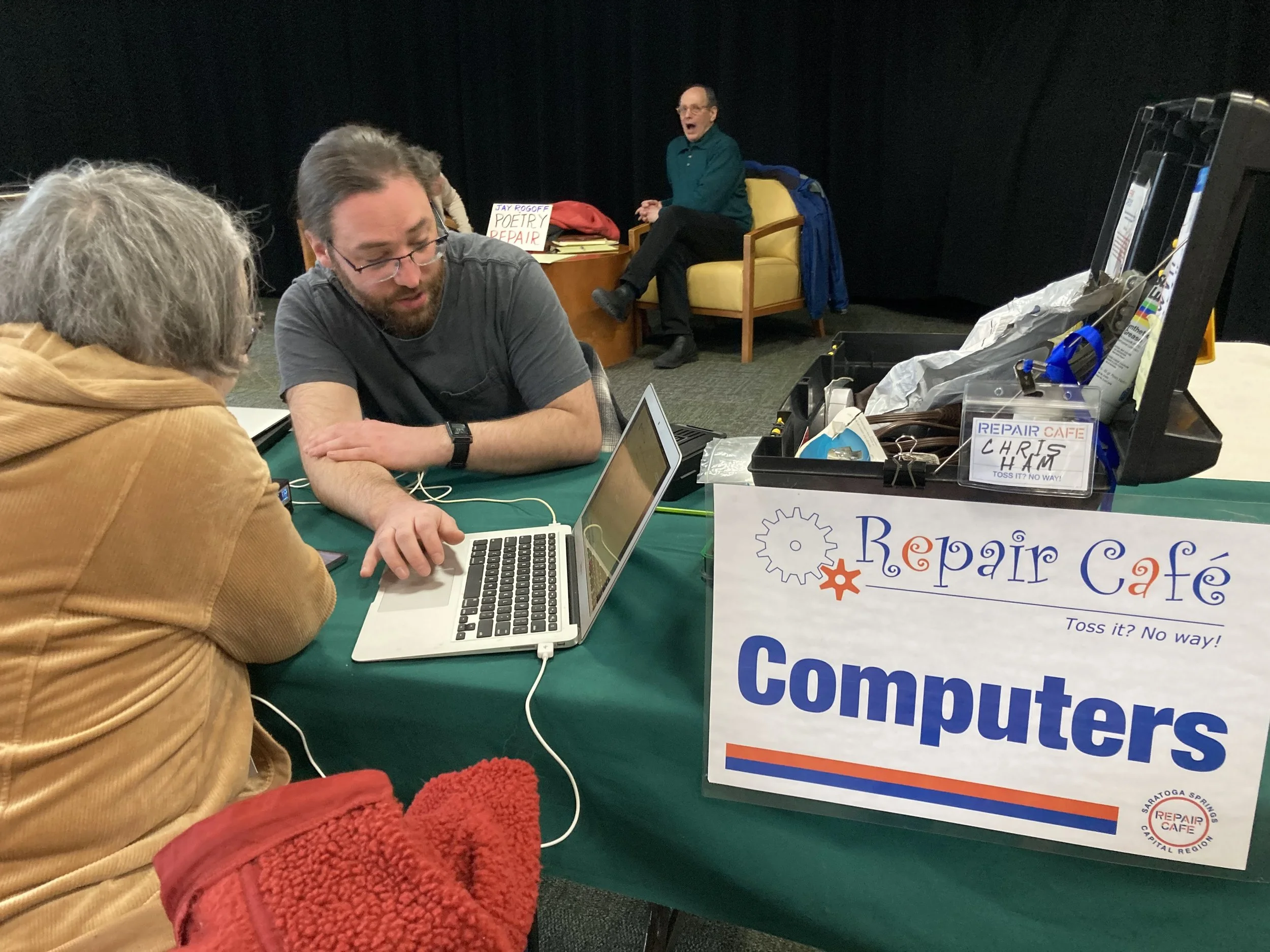 A man seated at a table helps a woman with her computer.