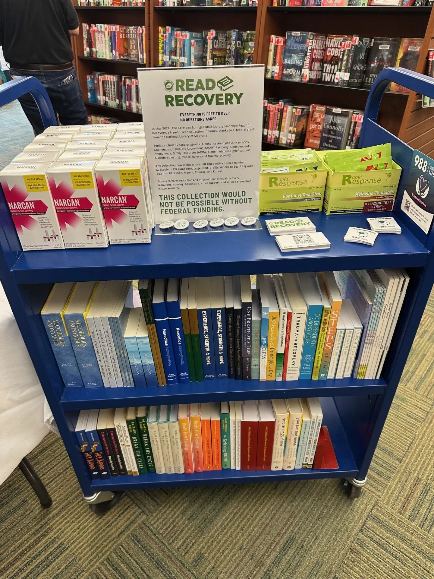 A bookcart with a sign saying "Read to Recovery" has narcan, fentanyl test strips, and books on addiction recovery.