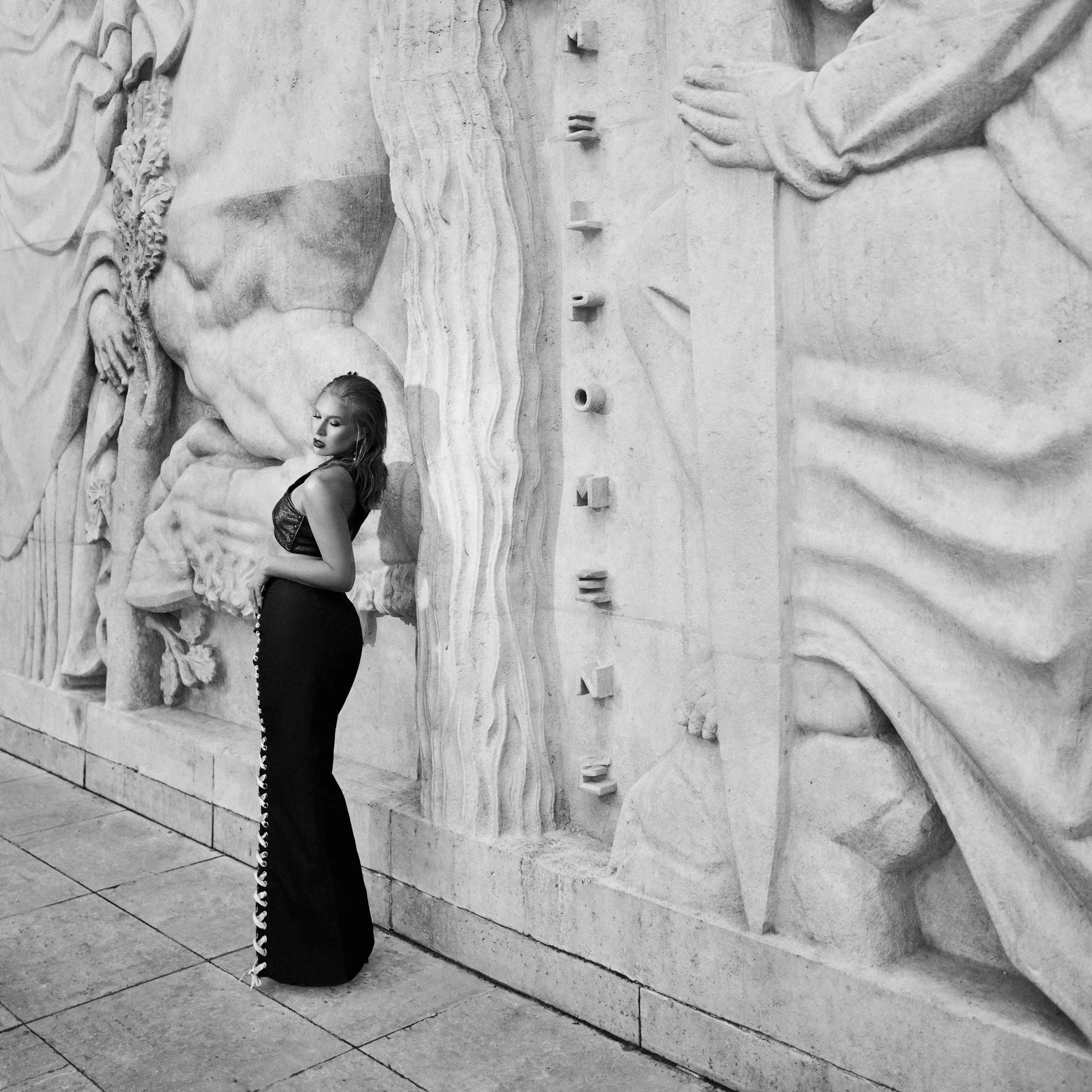 A woman wearing a long black dress with embellishments along the side, standing against a carved stone wall with relief sculptures.