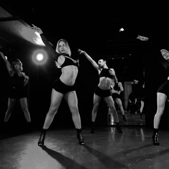 Group of young women dancing in a studio with dark walls and mirrored floor, some wearing crop tops and shorts.