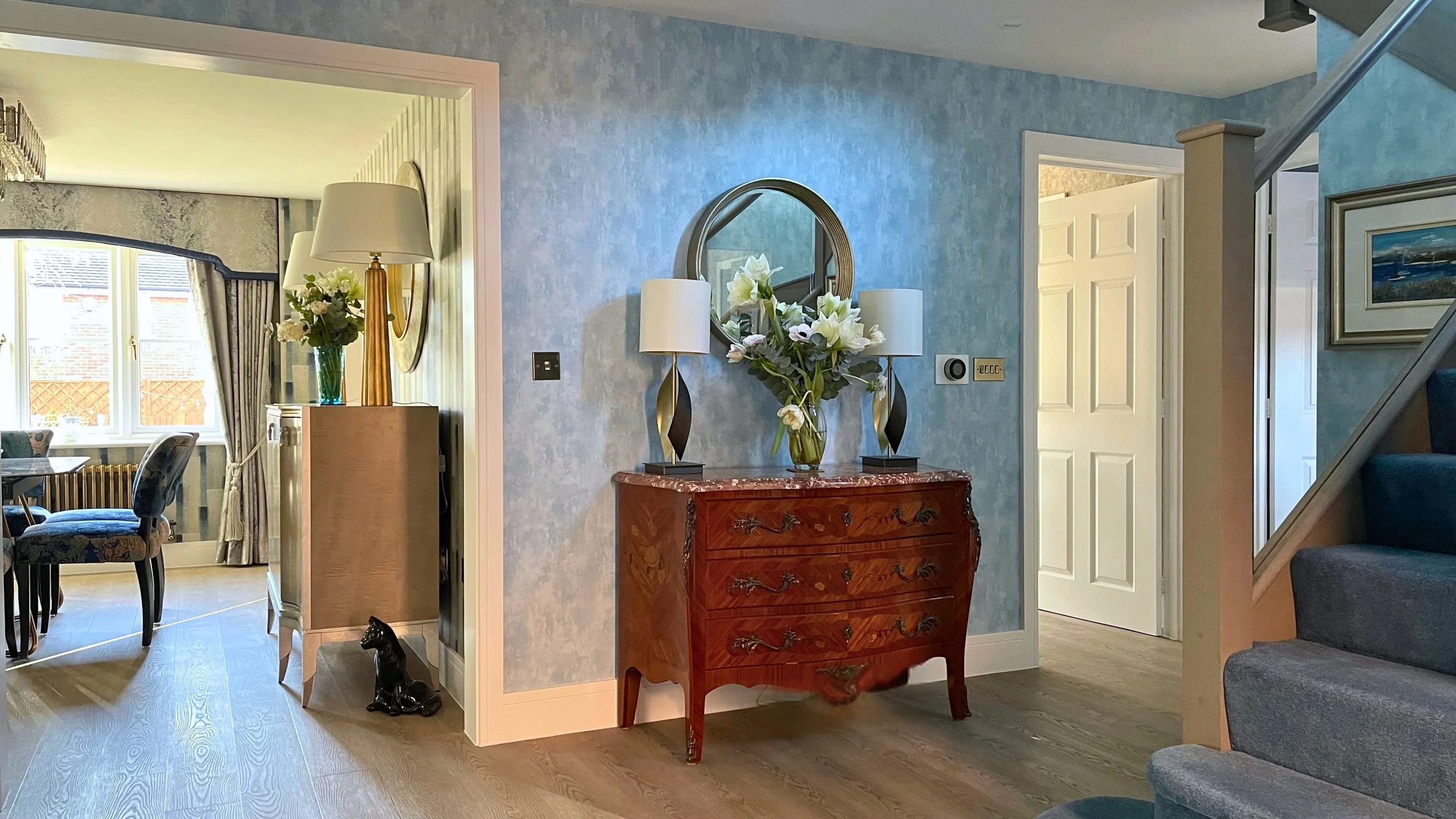 Blue wallpapered hallway with curved blue carpeted staircase and wooden flooring