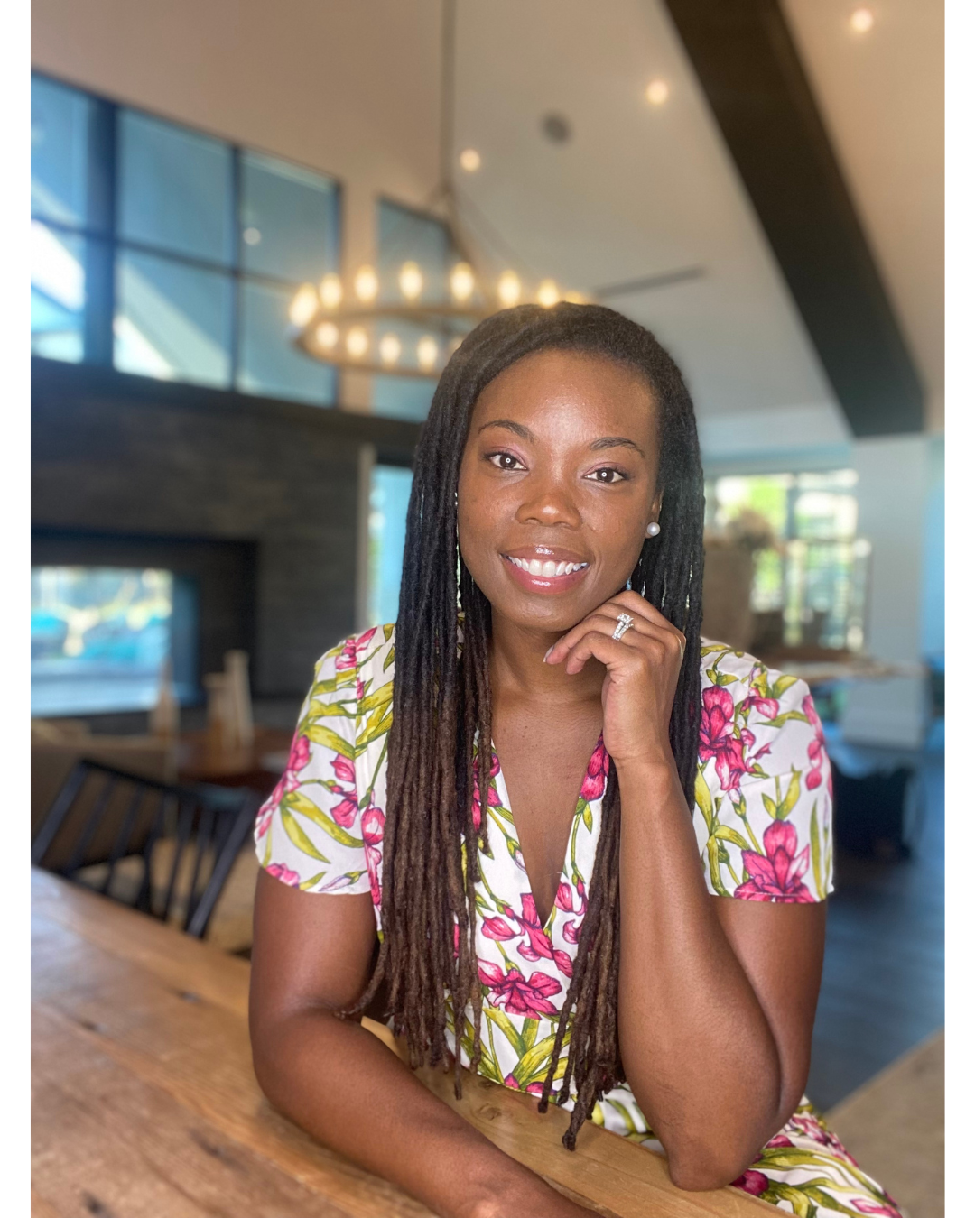 A woman with long dreadlocks and dark skin sitting at a wooden table in a modern restaurant, smiling, wearing a floral dress with pink flowers and green leaves, holding her chin with her right hand, with large windows and a chandelier in the background.