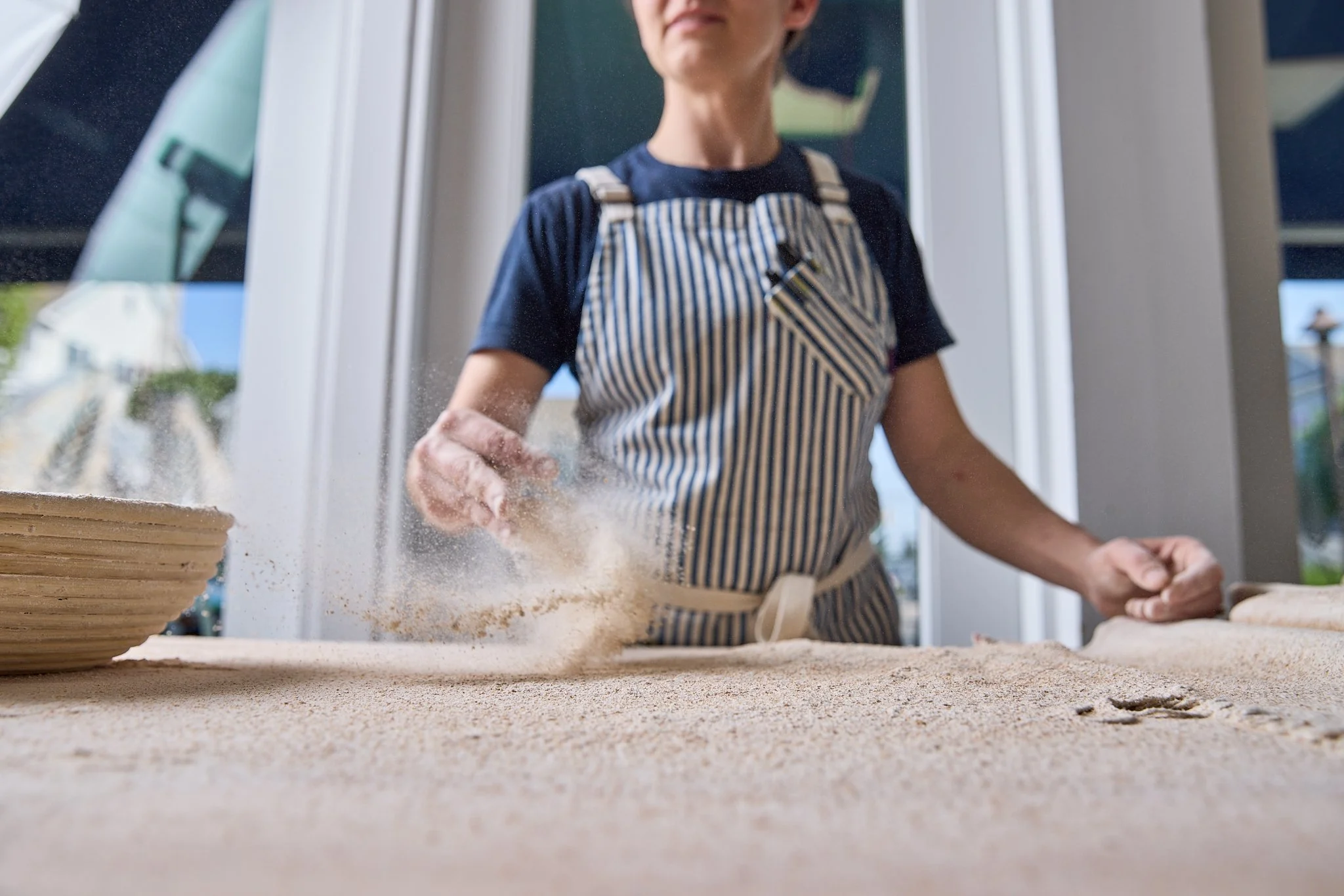Person wearing a striped apron pounding dough on a wooden surface, dusting flour in a well-lit kitchen with a window and houses outside.