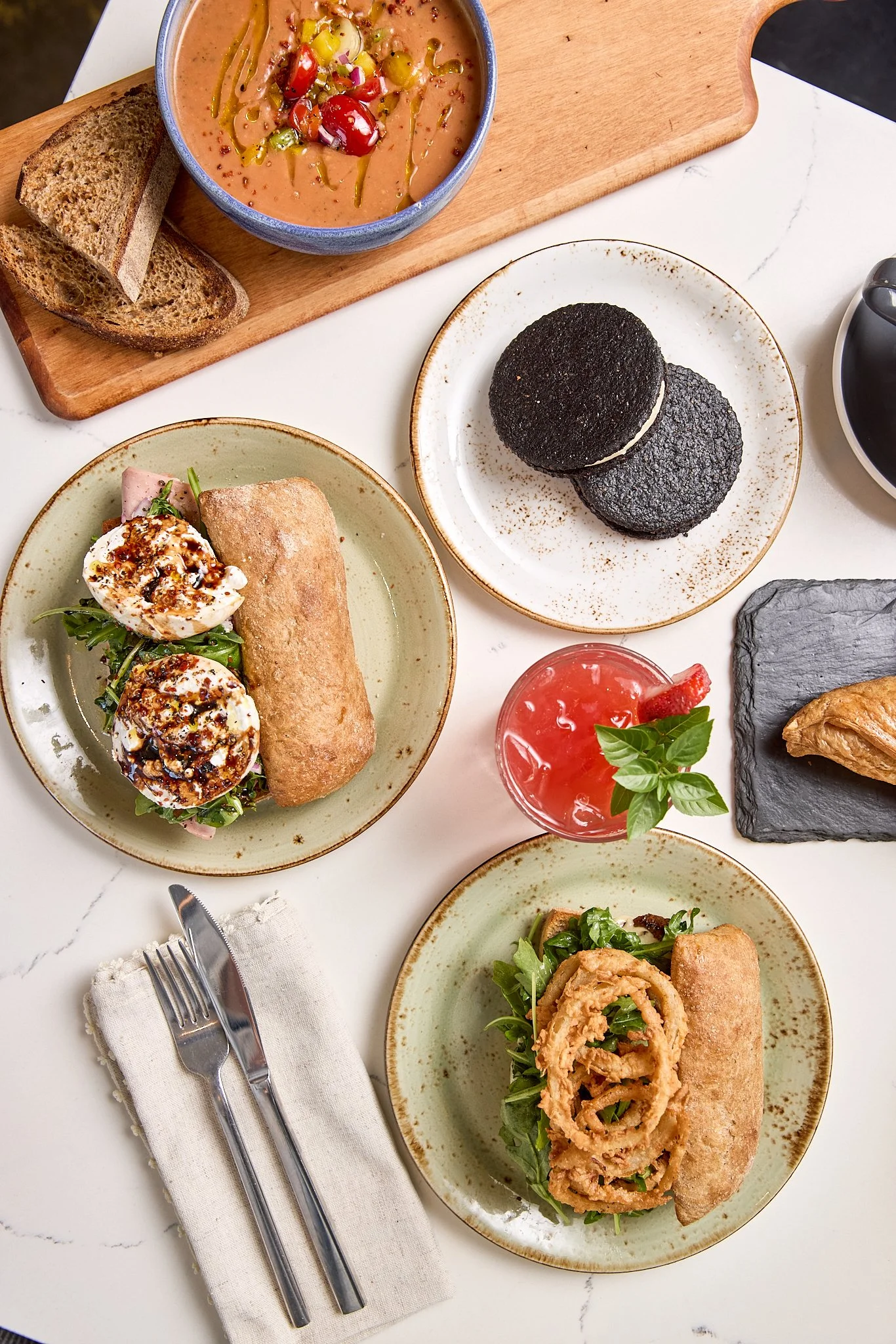 A table with two spring roll sandwiches, a bowl of mixed bean soup, a black Oreo cookie cream sandwich, a glass of pink cocktail garnished with basil, and a plate with fried onion rings and a spring roll, with cutlery on a cloth napkin.