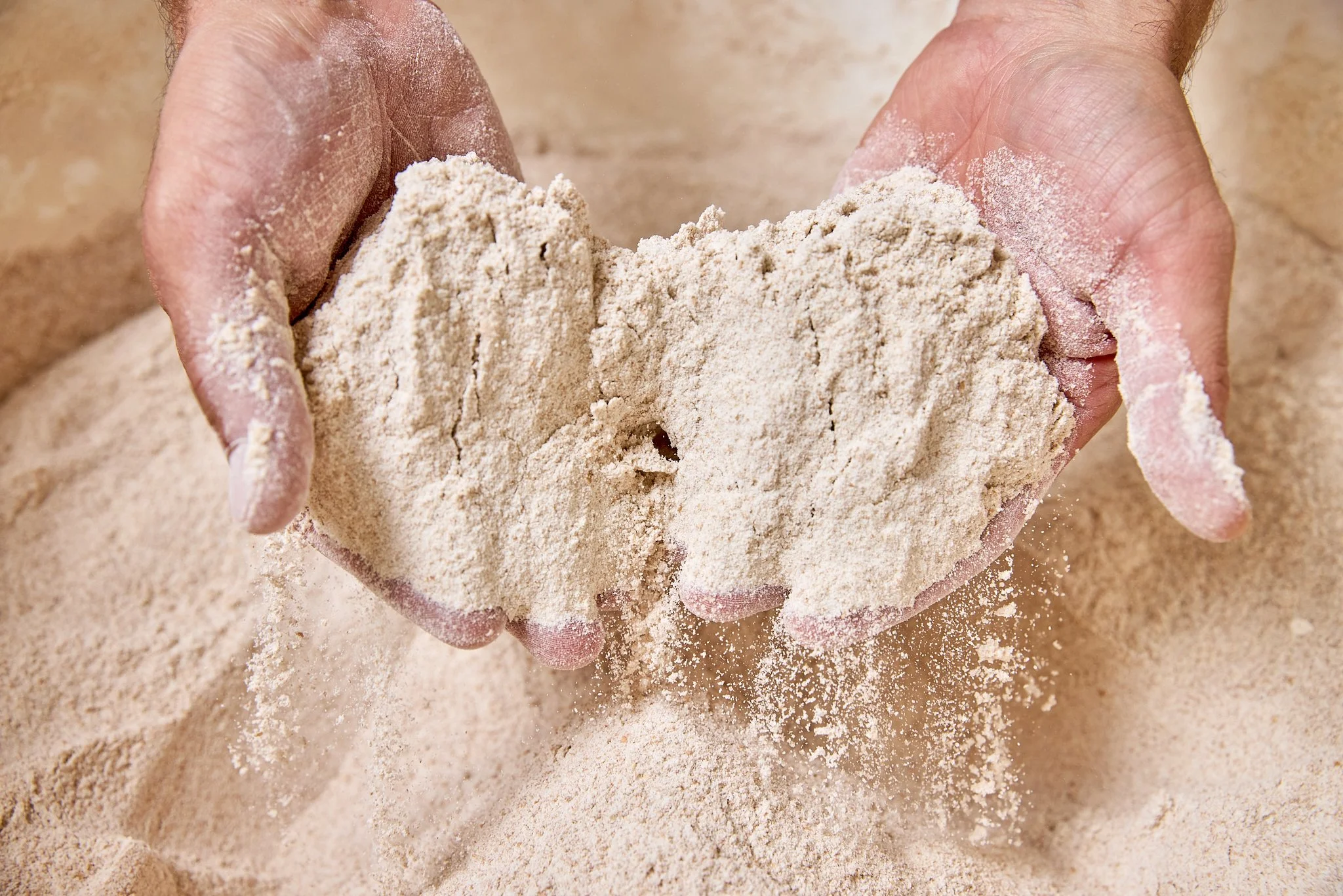 A person holding a handful of white or light beige flour over a surface covered with more flour.