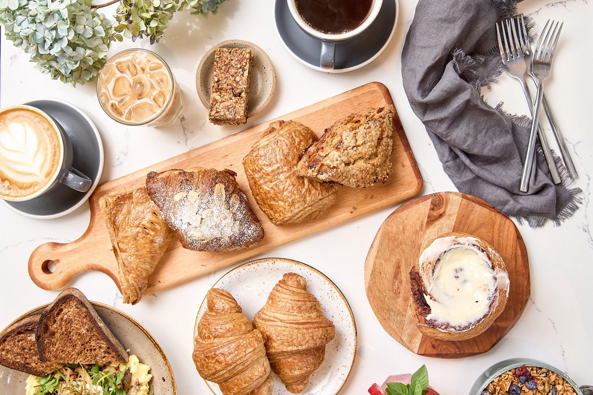 Assorted breakfast items on a marble table, including croissants, muffins, cake slices, a latte, coffee, iced coffee, and granola.