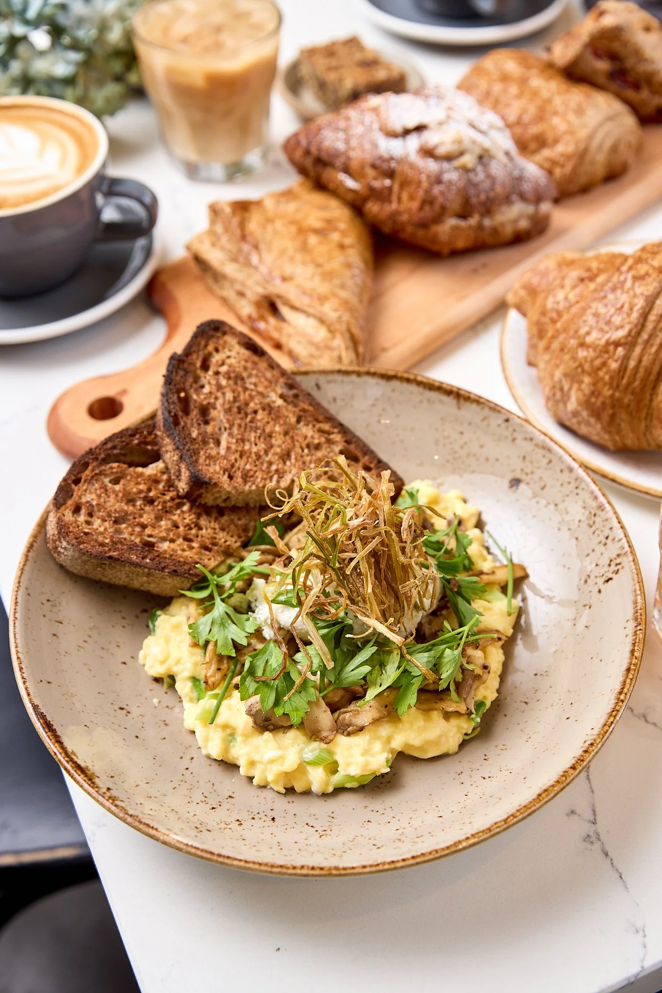 A bowl of scrambled eggs topped with herbs and sautéed mushrooms, served with slices of toasted bread, on a table with various baked goods and coffee.