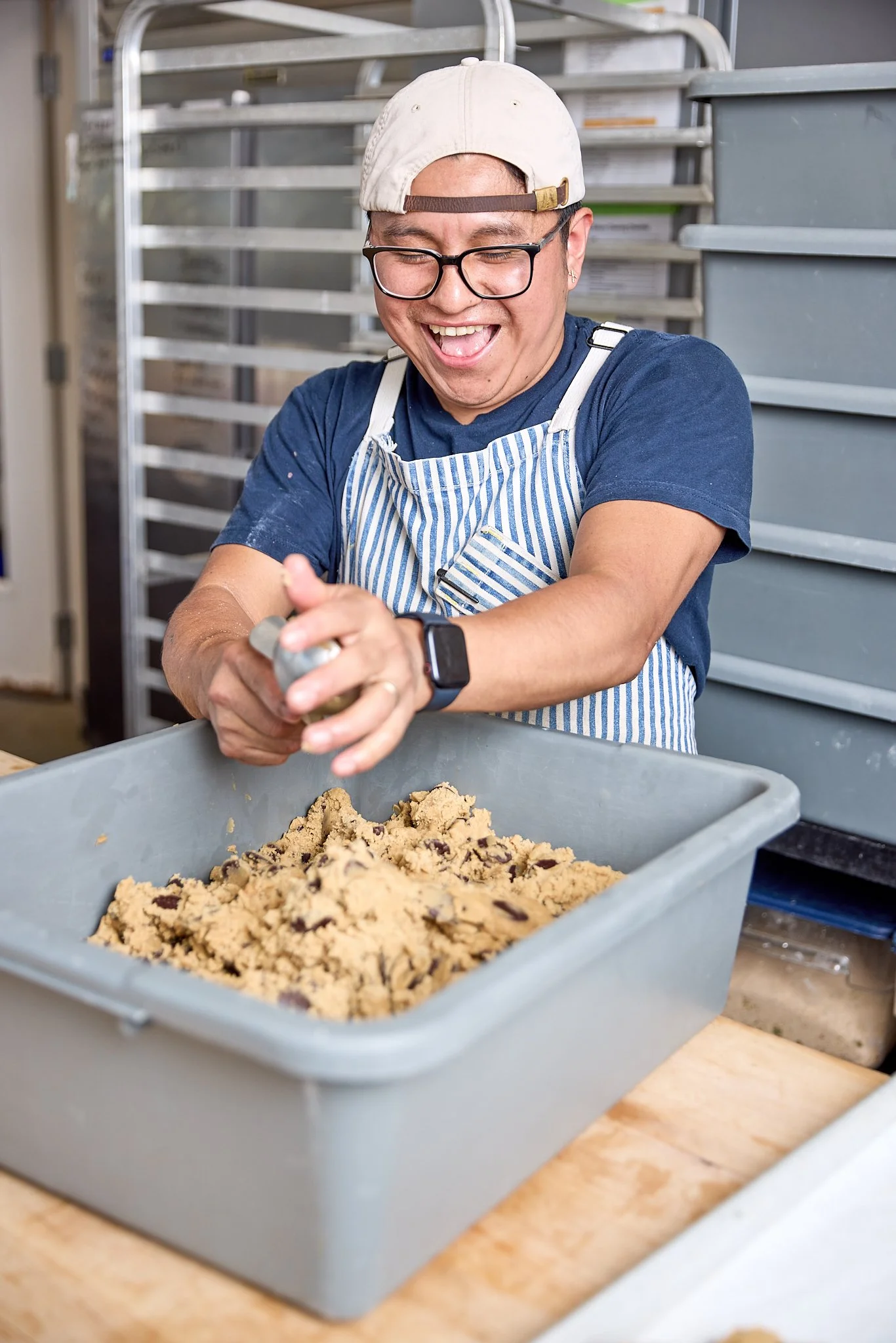 A person smiling happily while sprinkling salt onto chocolate chip cookie dough in a gray plastic container in a bakery or commercial kitchen setting.