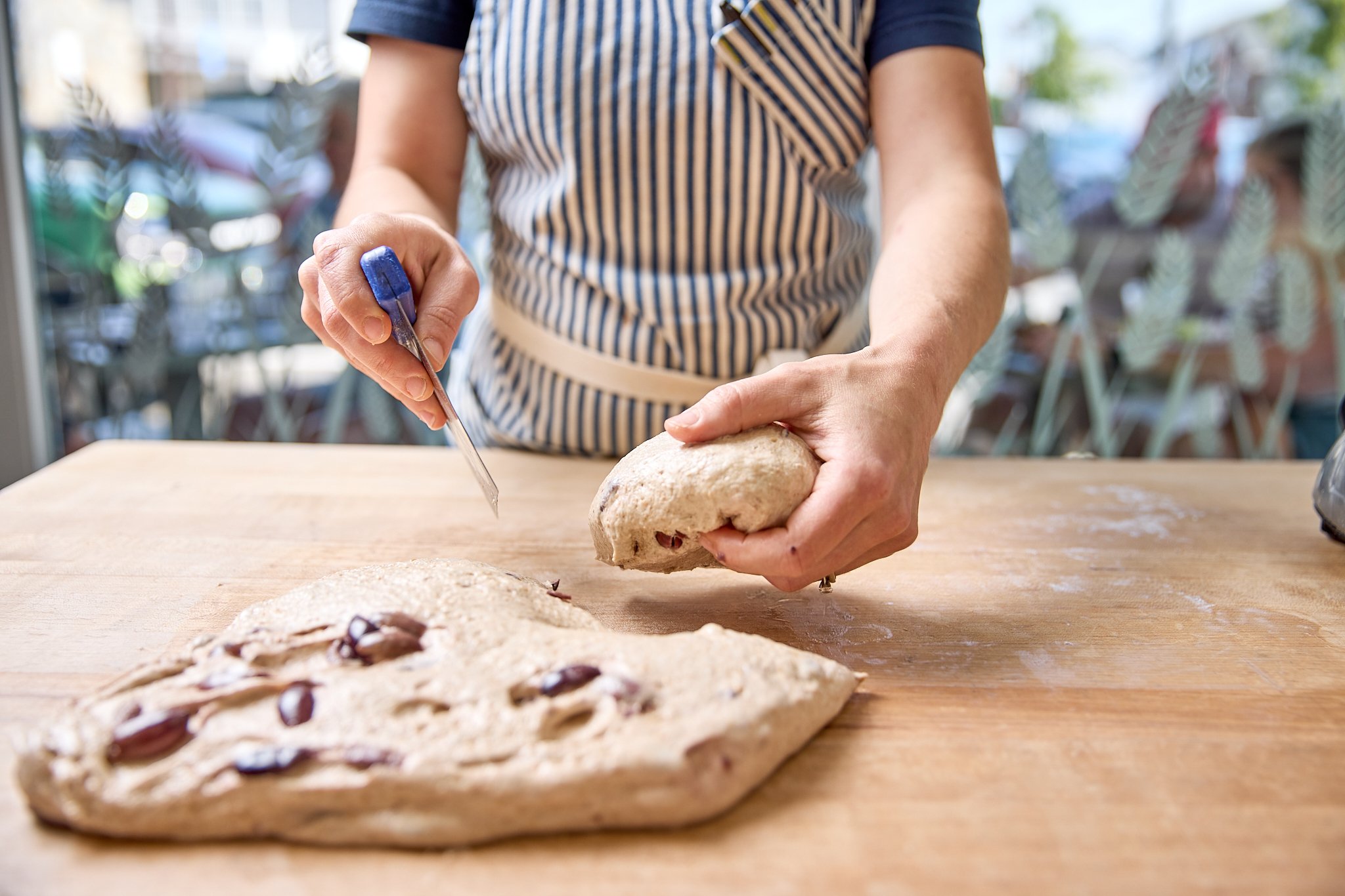 Person in a striped apron is shaping pizza dough on a wooden table with a knife in one hand and the dough in the other. A piece of folded pizza with toppings is on the table.