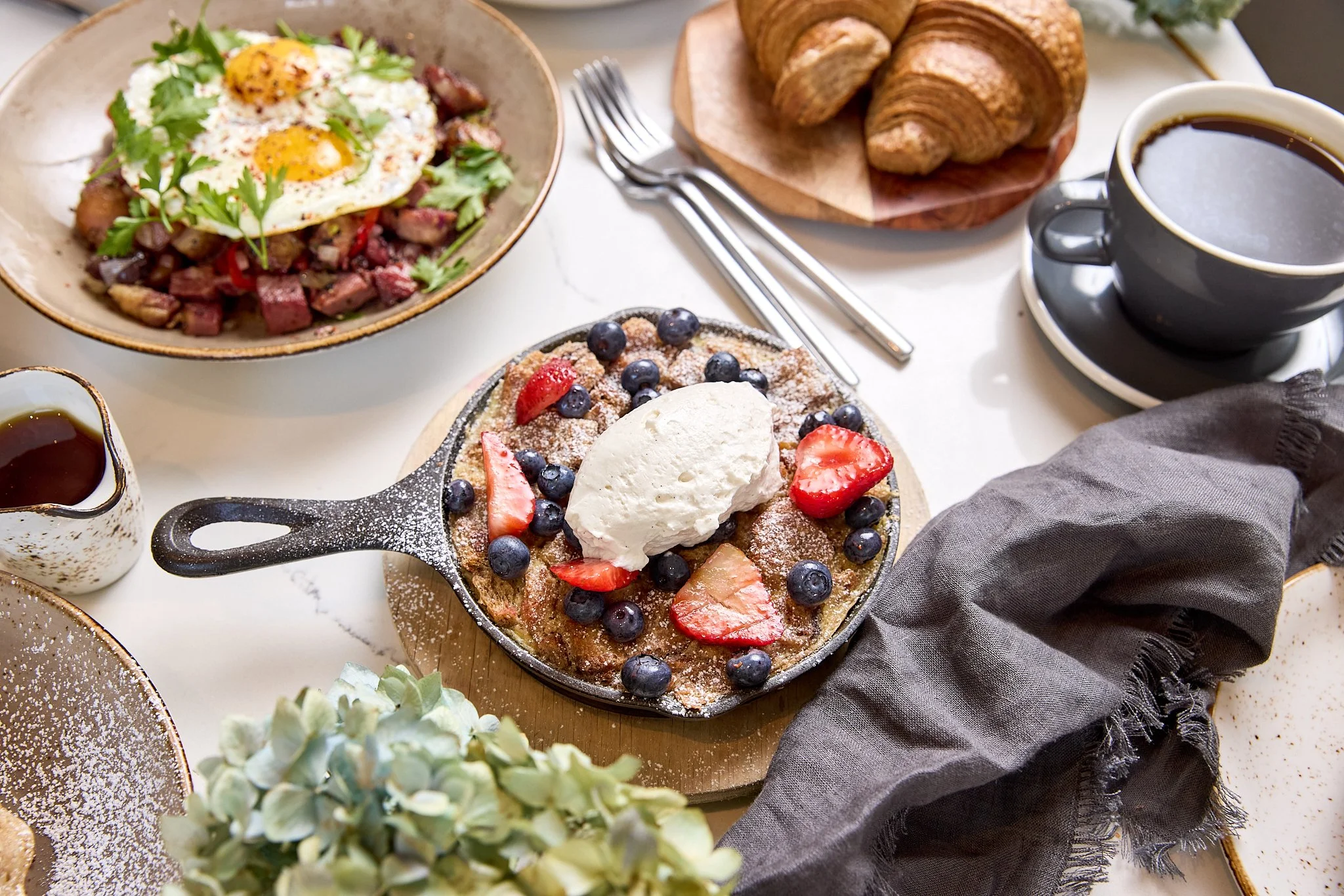 Breakfast scene featuring a skillet with strawberries, blueberries, whipped cream, and powdered sugar, a bowl of hash with eggs, bacon, toast, a cup of coffee, and croissants on a table.