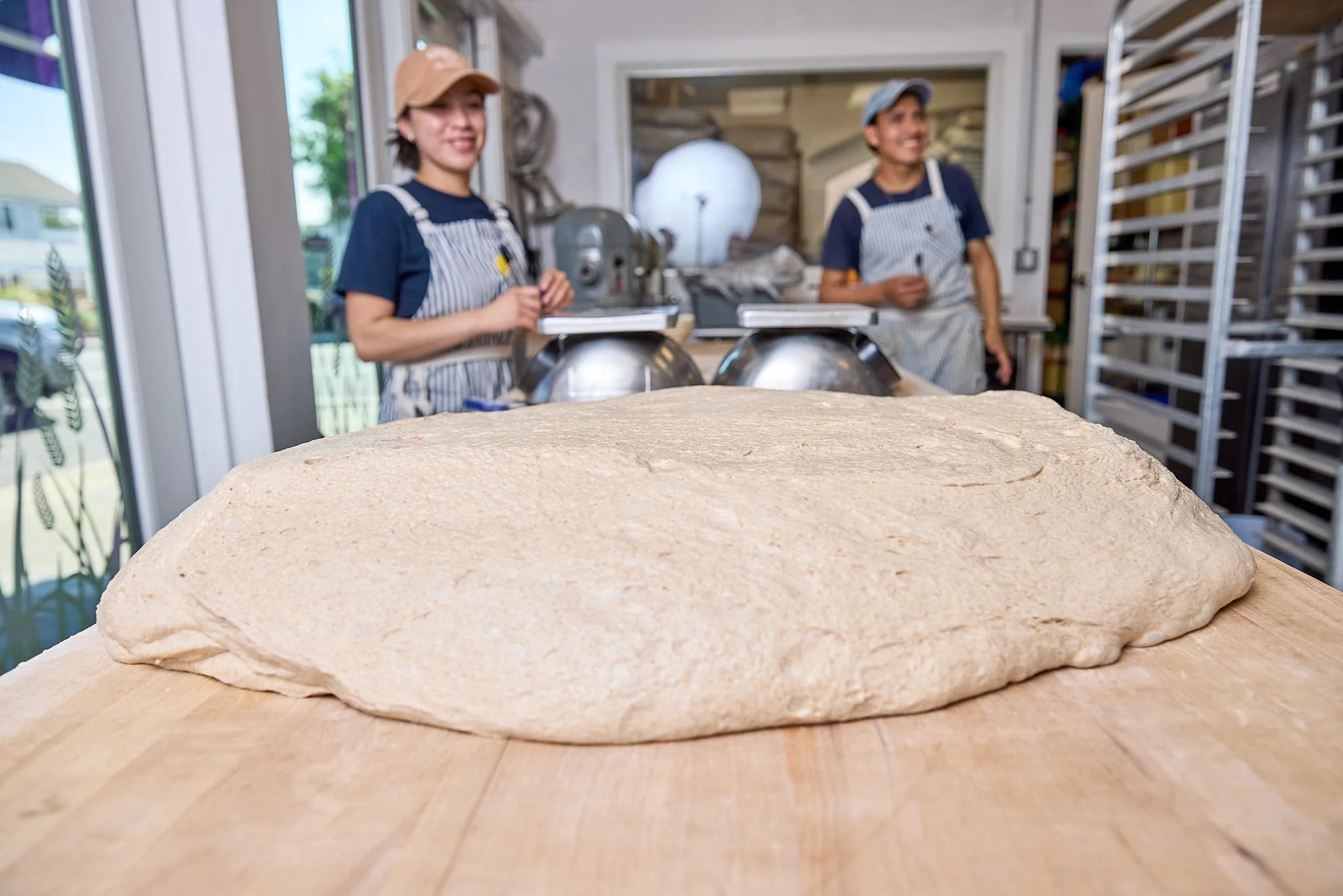 Raw pizza dough on a wooden surface with two bakers in aprons and hats smiling in the background.