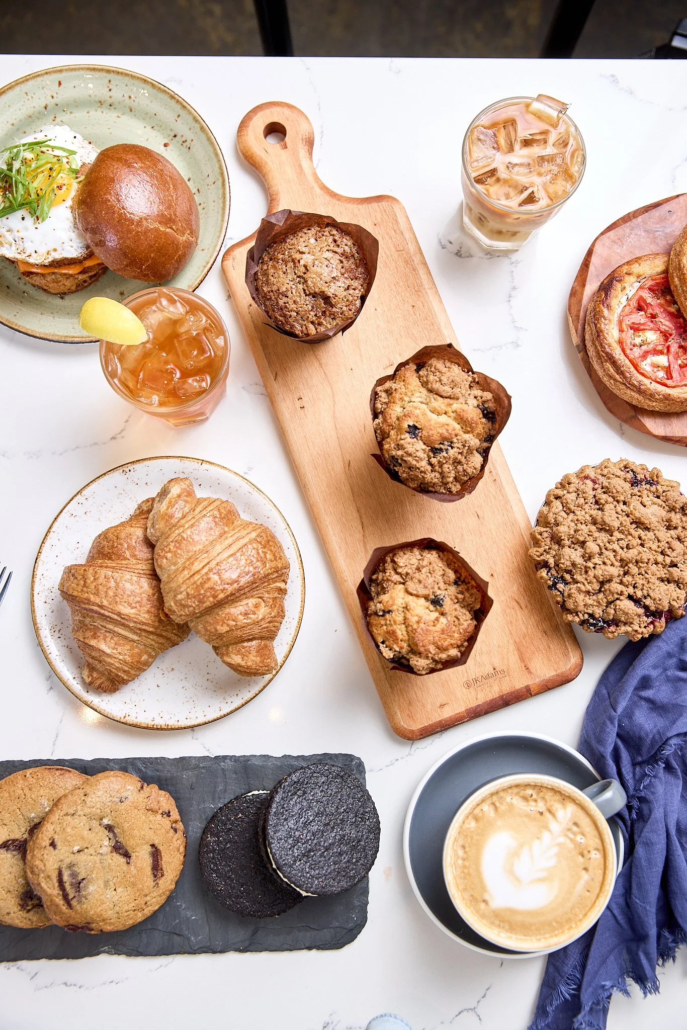 A top-down view of a breakfast spread on a white marble table, featuring croissants, cookies, muffins, coffee with latte art, iced drinks, and small plates of sandwiches and pastries.