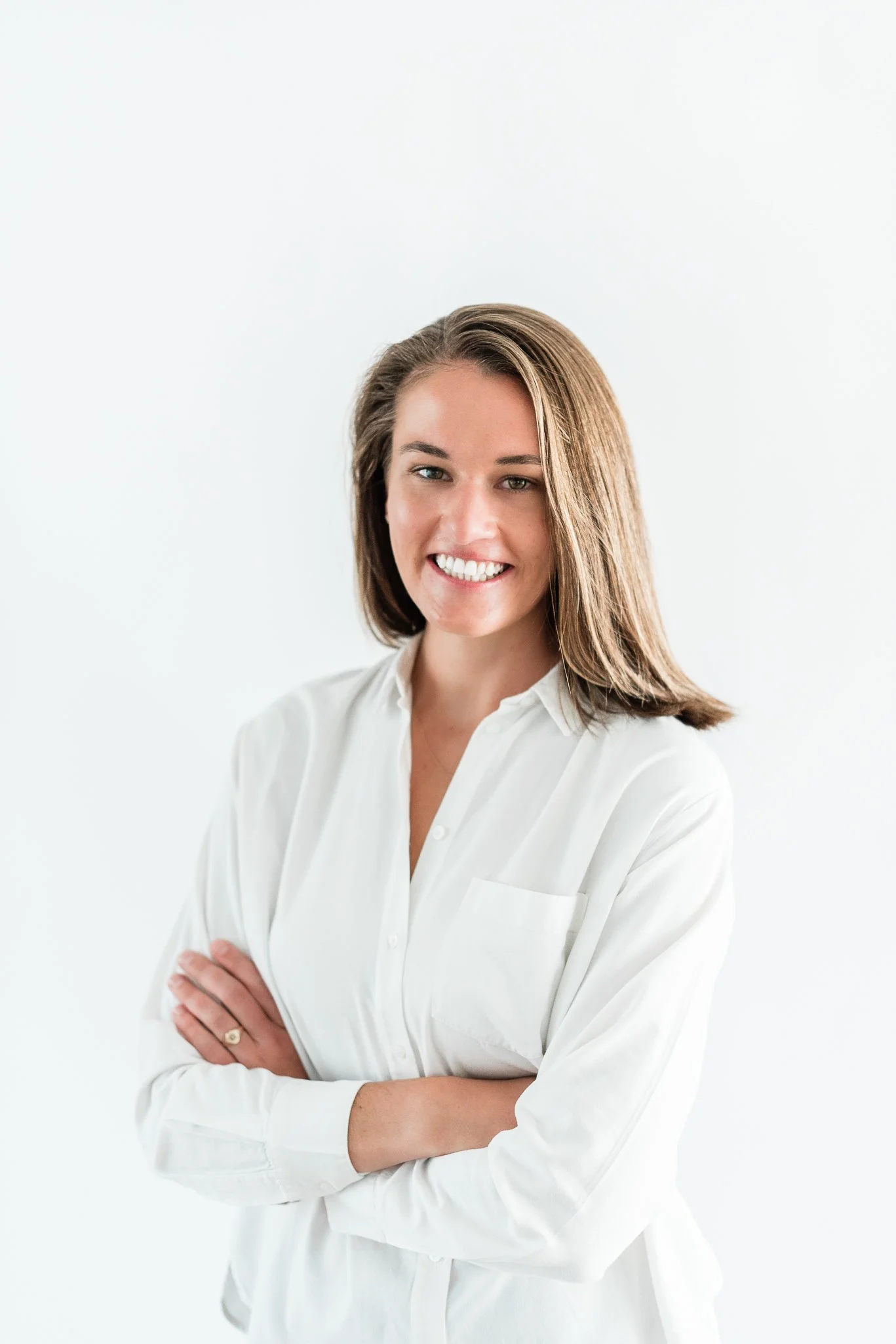 A woman with shoulder-length brown hair smiling, wearing a white button-up shirt, standing against a plain white background.