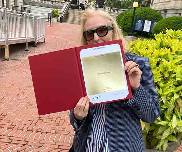 A woman with blonde hair and glasses holding up a red folder or certificate, partially covering her face, outdoors in front of green shrubs and yellow plants.