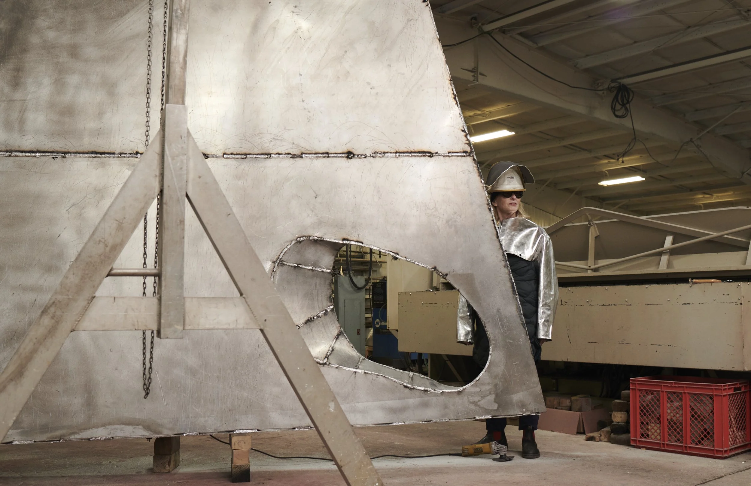 A person wearing protective welding gear, including a helmet, sunglasses, and a silver apron, stands beside a large metal structure in an industrial workshop.
