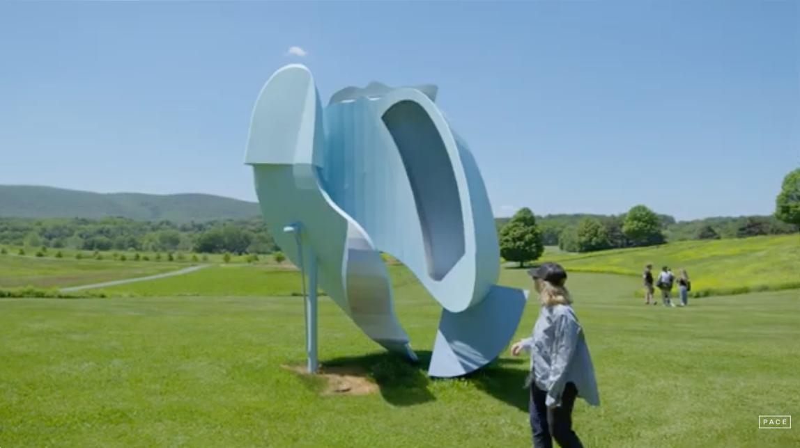 A large abstract metallic sculpture on a grassy field with people walking around, trees, hills, and a clear blue sky in the background.