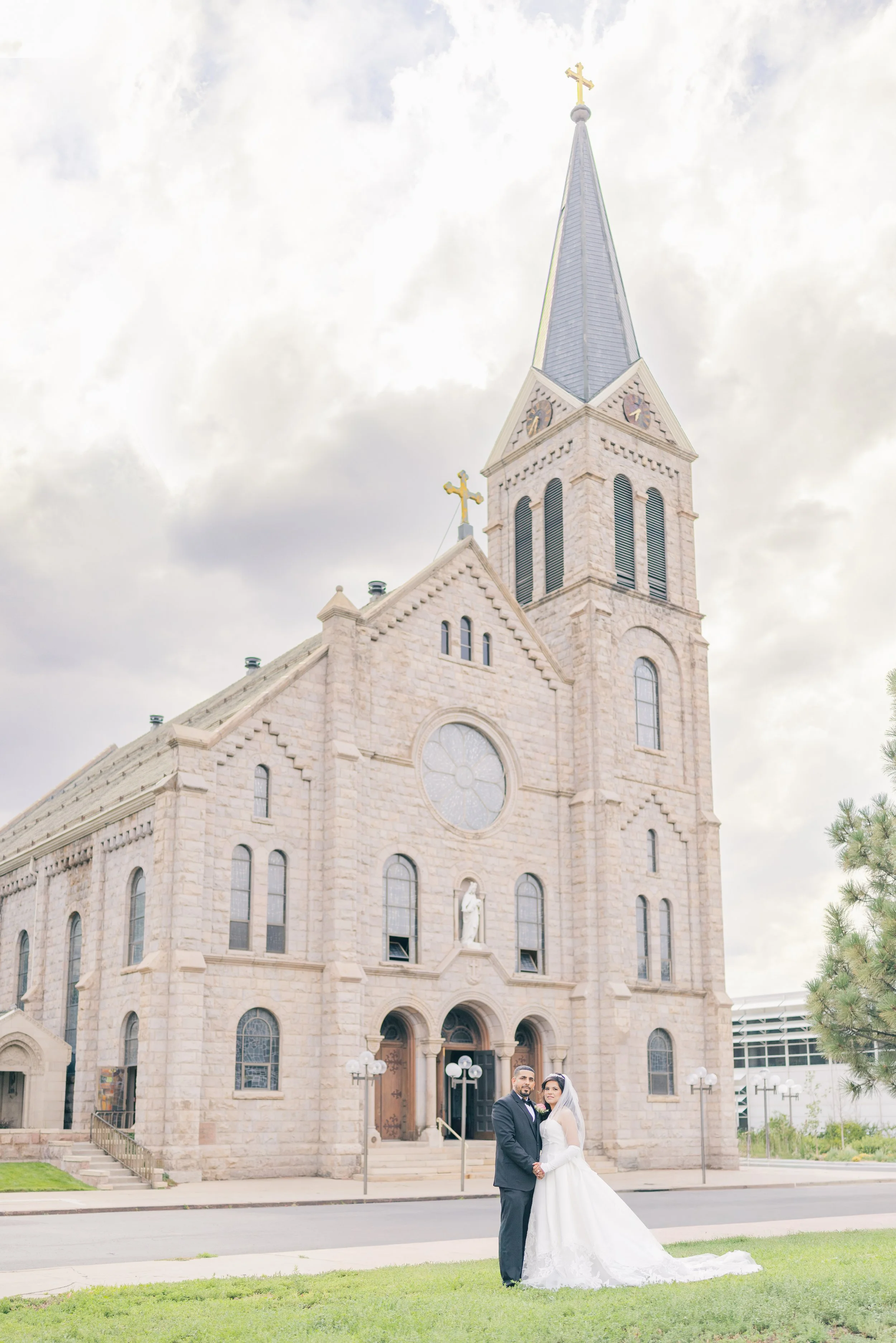 A newly married couple standing outside a stone church, holding hands, with the bride wearing a white wedding dress and the groom in a dark suit. The church features a tall steeple topped with a cross and a statue above the main entrance. Wedding