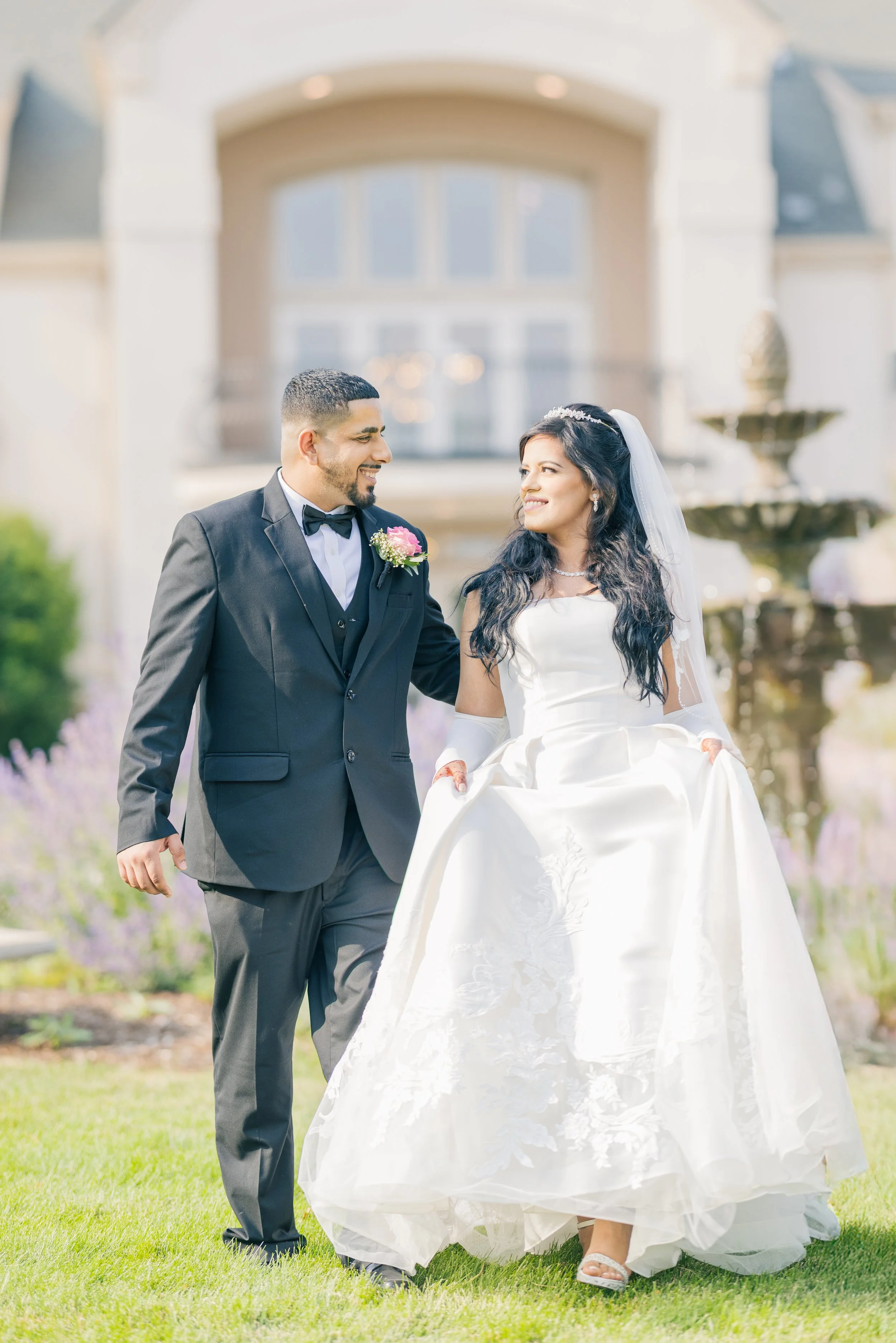 A bride and groom walking on a lawn during their wedding, with the bride wearing a white wedding gown and veil, and the groom in a dark tuxedo with a bow tie, smiling at each other. Wedding
