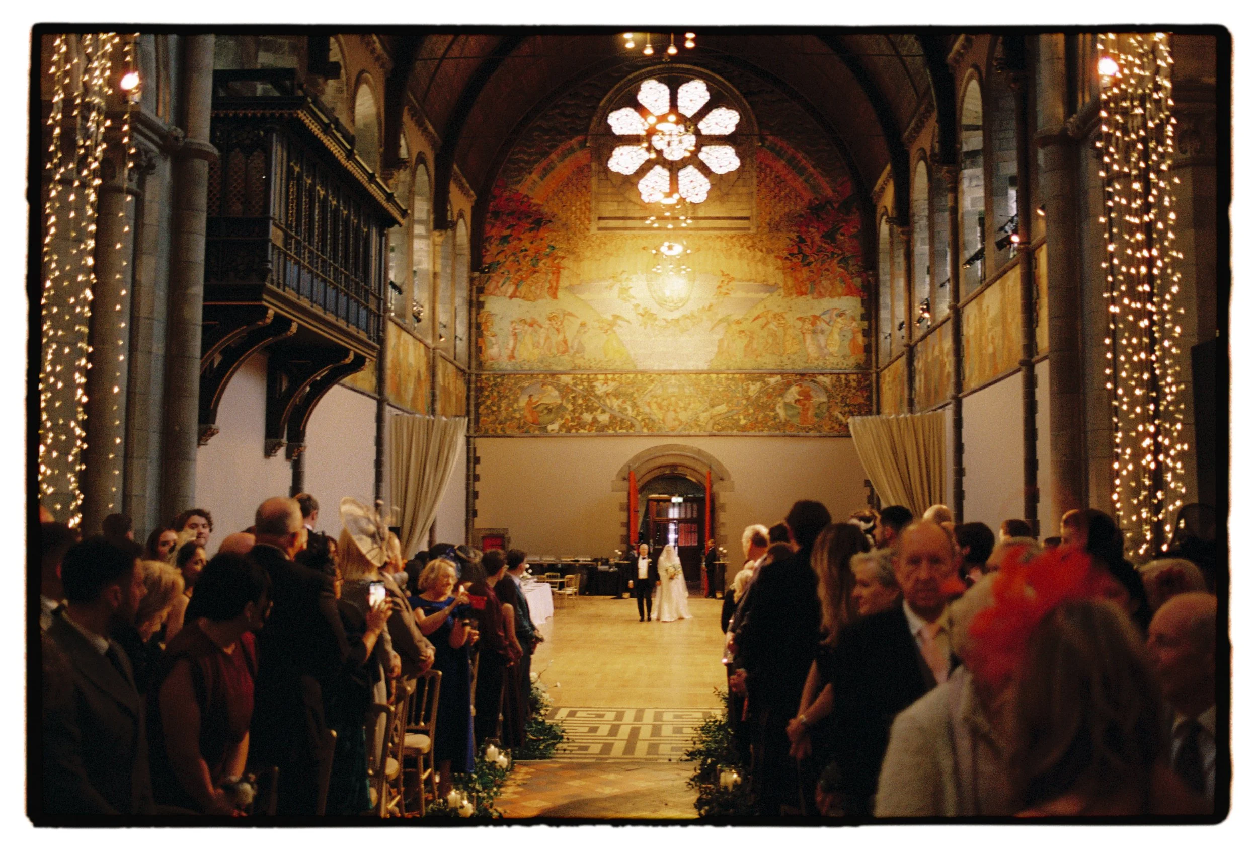 Bride and father walk into ceremony at Mansfield Traquair in Edinburgh through big red doors into a large cathedral like space with painted murals. Wedding guests stand in the foreground either sid of the aisle watching the bride and her father