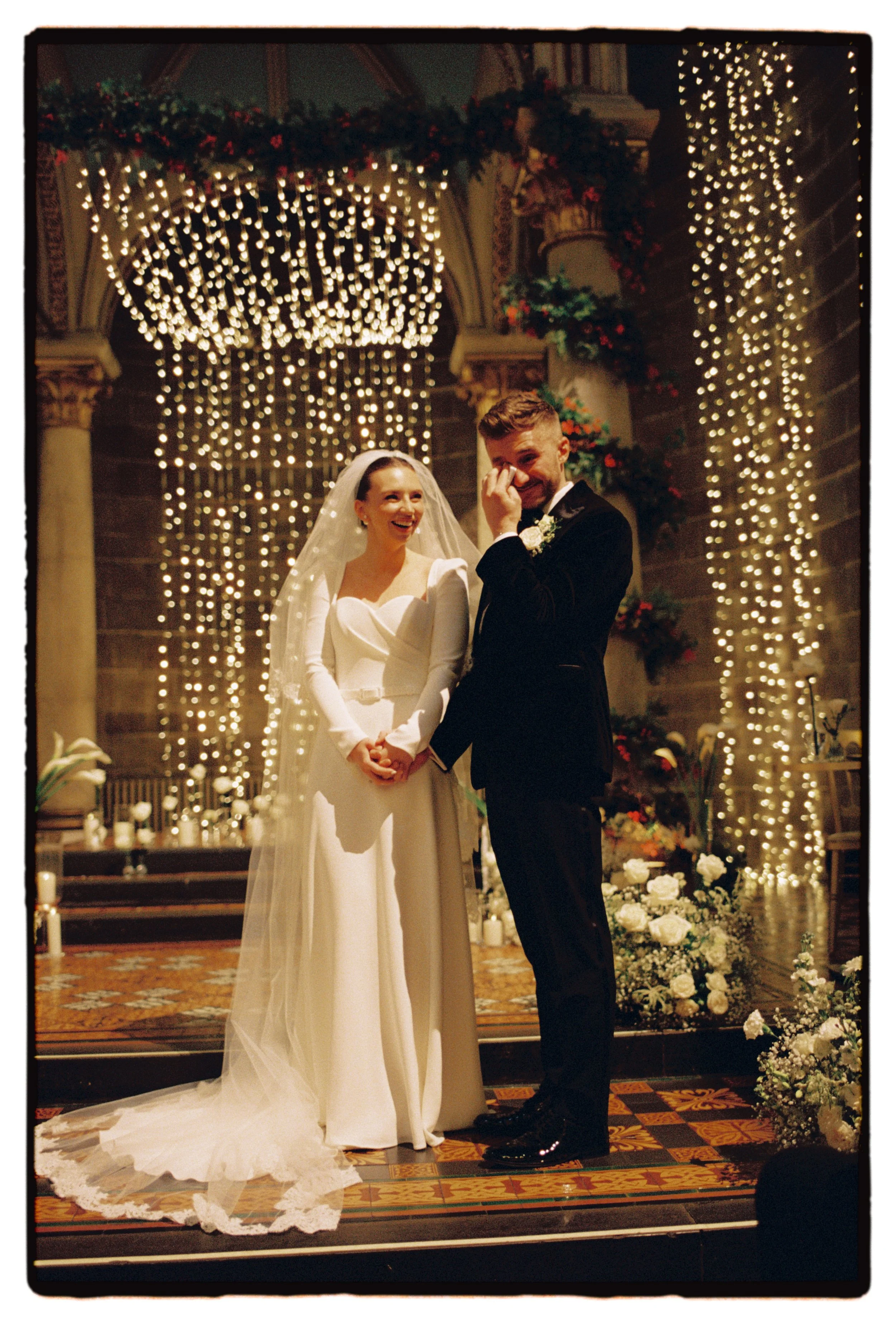 Bride and groom stood at the front of their indoor ceremony in a old cathedral with lots of fairy lights and white roses surrounding them. The groom wipes a tear from his face and the bride smiles