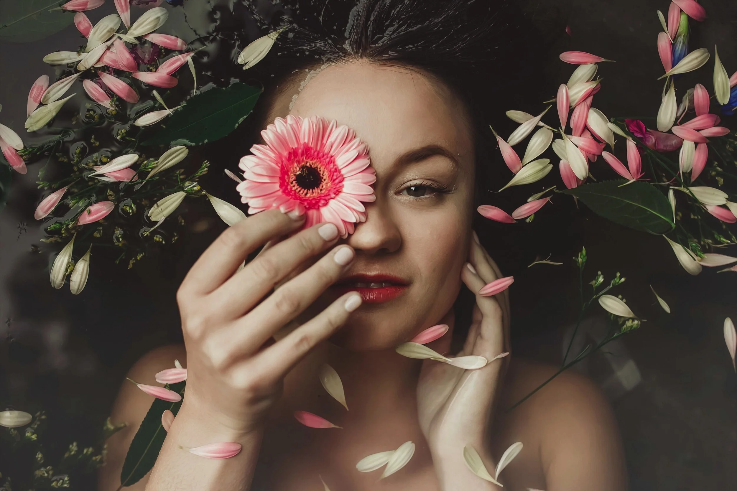 A woman lying in water surrounded by pink and white flower petals, holding a pink flower in front of one eye.